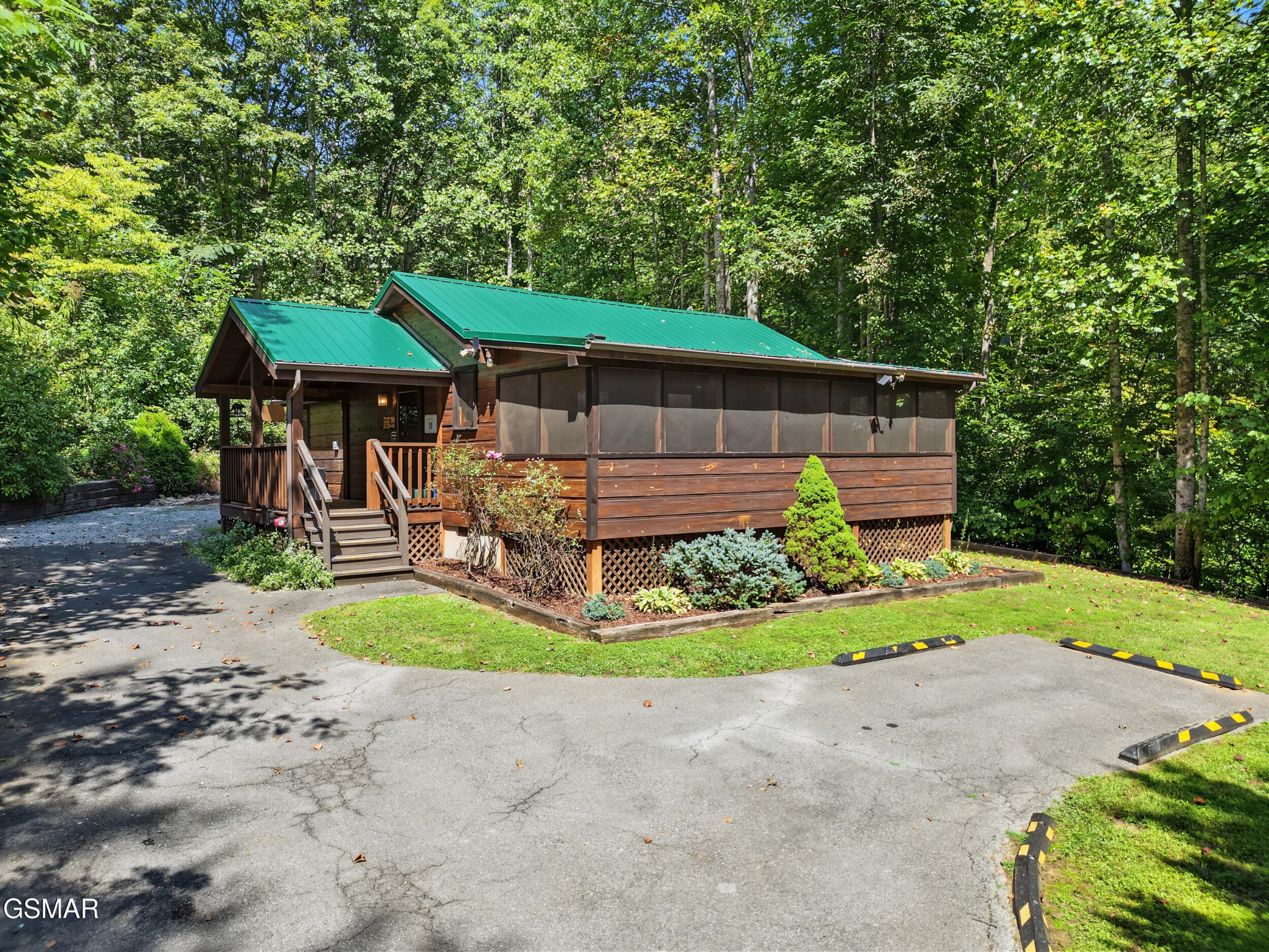 Wood cabin with a green metal roof and screened porch, surrounded by trees beside a paved parking area.