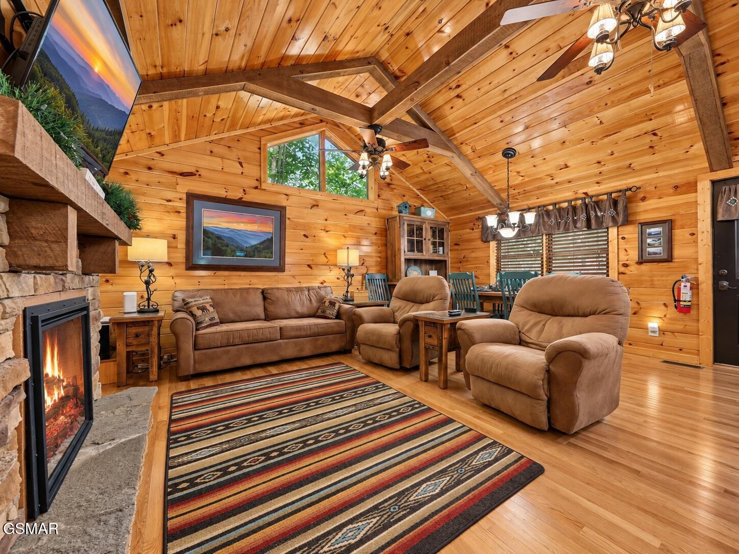 Cabin living room with vaulted pine ceiling, stone fireplace, leather seating, and a colorful striped rug.