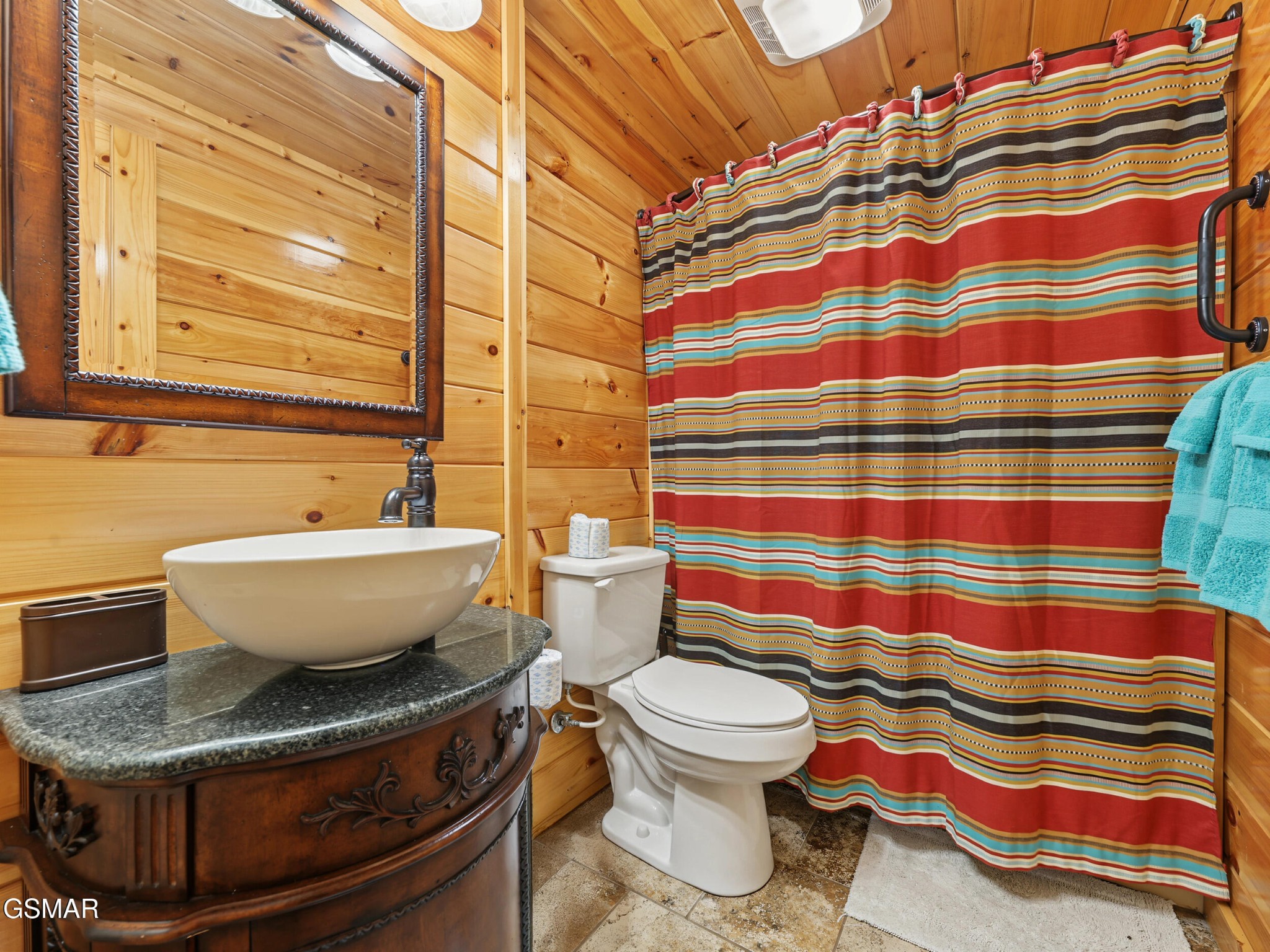 Wood-paneled bathroom with a carved vanity, vessel sink, toilet, and bold striped shower curtain.