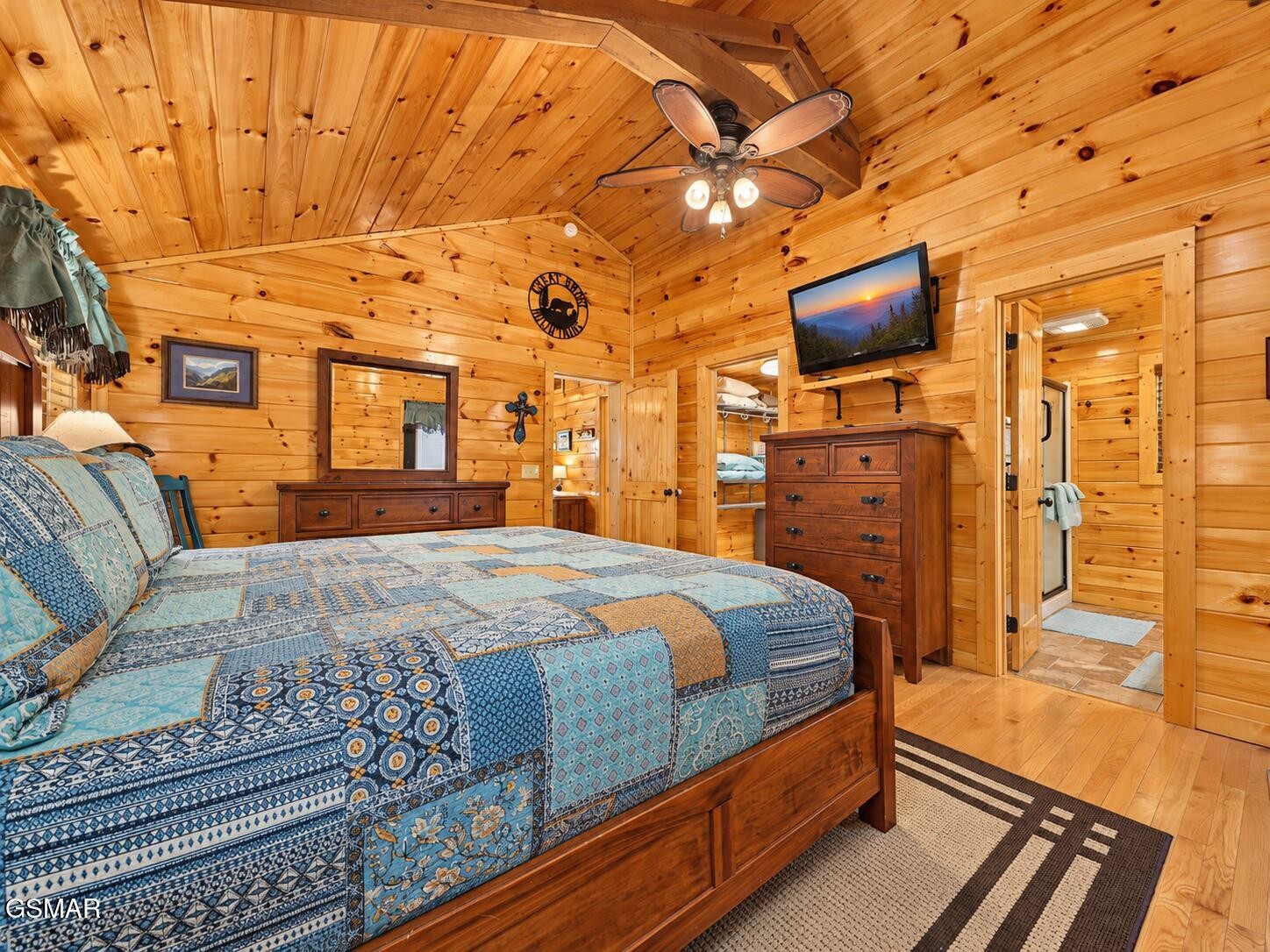 Cabin bedroom with knotty-pine walls, a blue patchwork bedspread, wooden dressers, and an open doorway.
