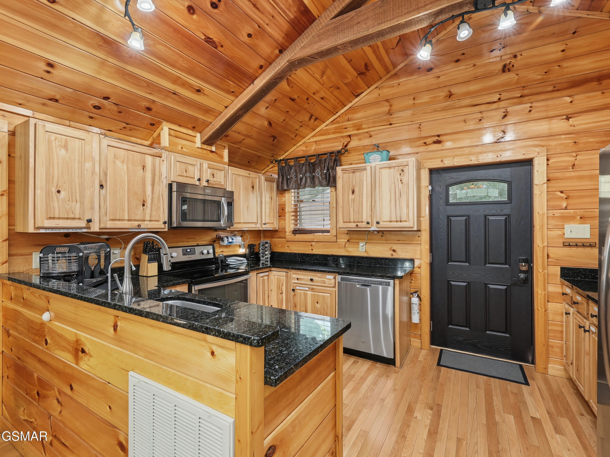 Pine-paneled cabin kitchen with vaulted ceiling, black granite counters, stainless appliances, and a black entry door.