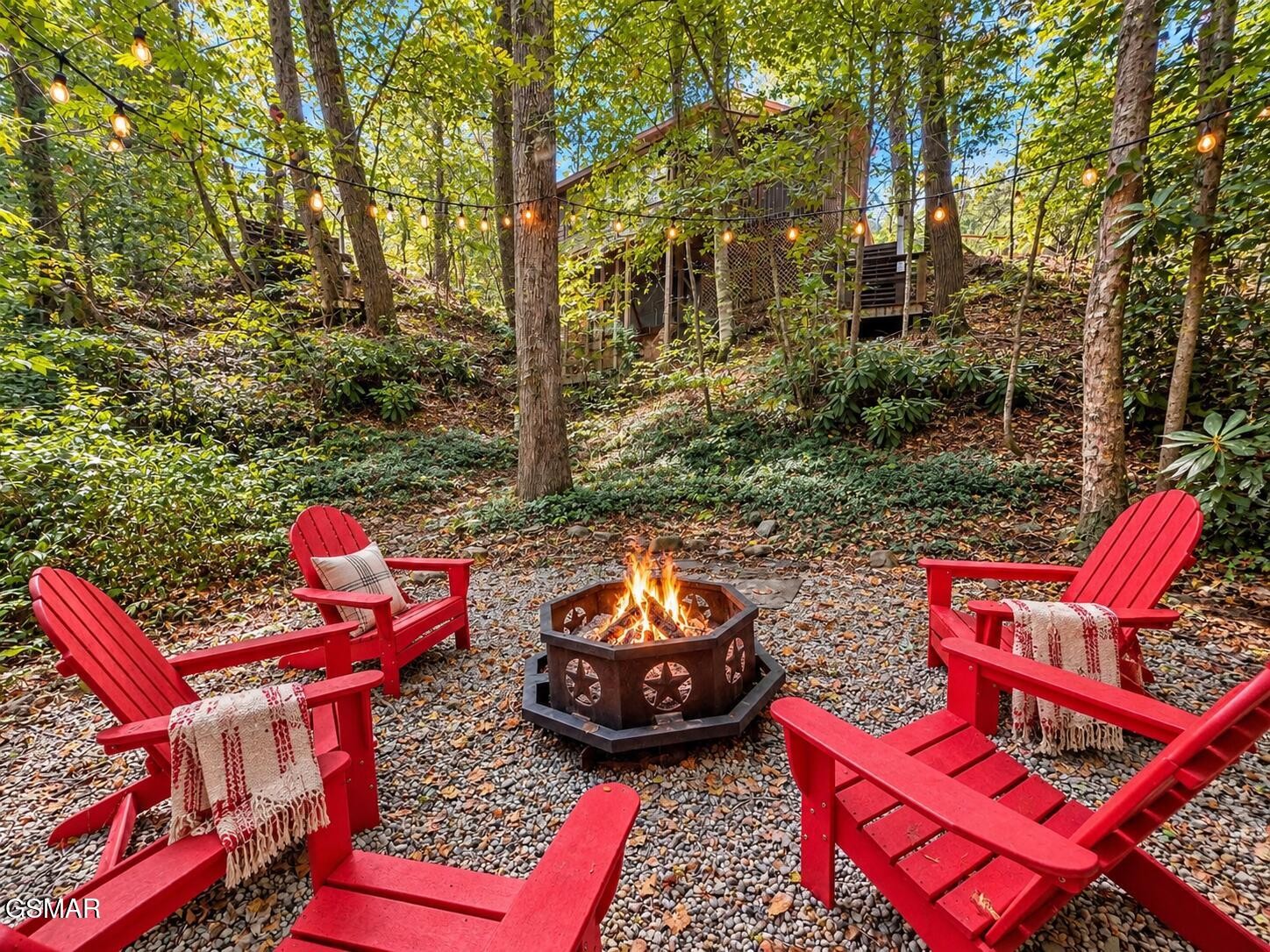 Wooded outdoor seating area with red Adirondack chairs, a fire pit, and overhead string lights.