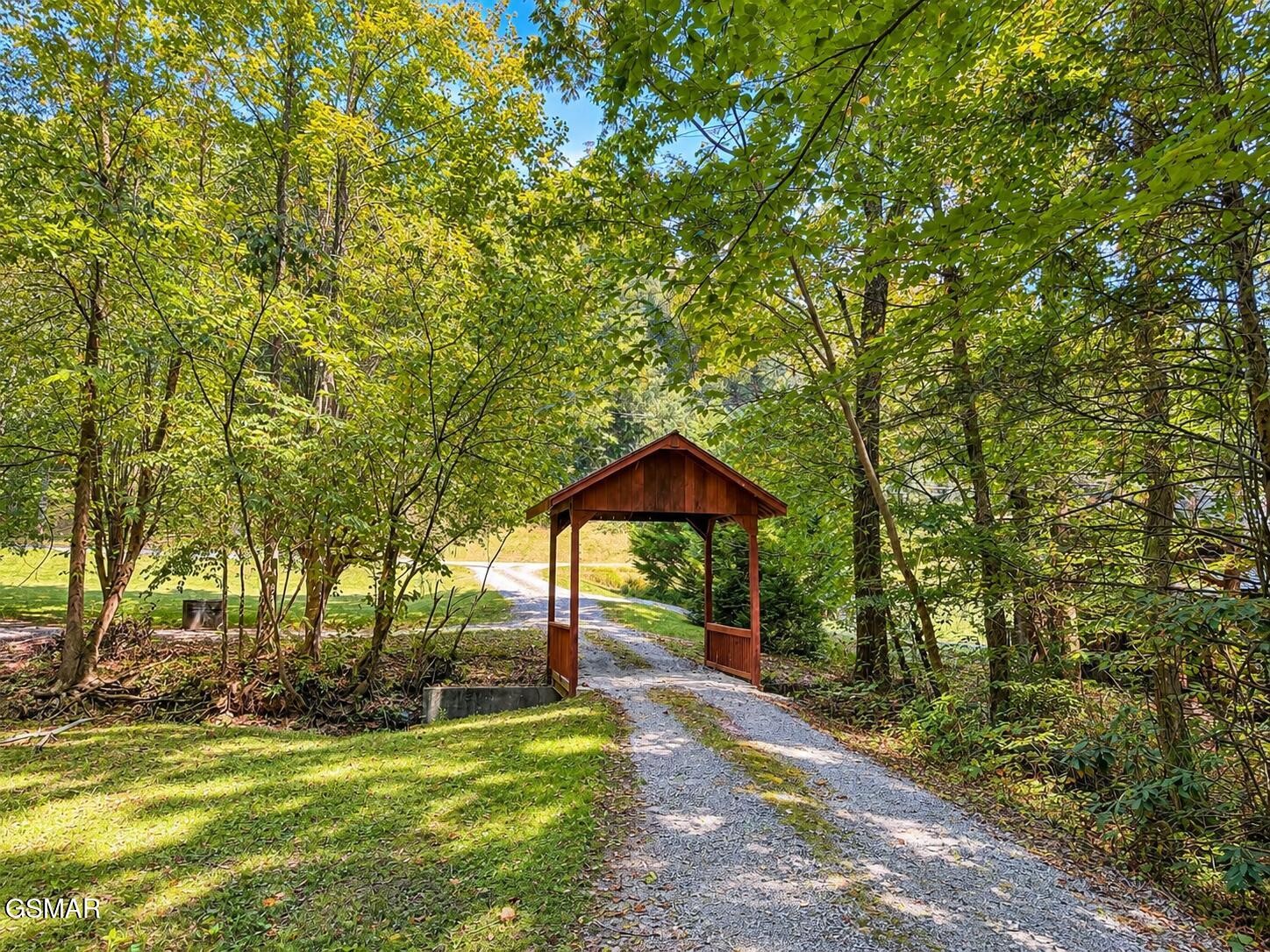 Gravel driveway passing beneath a wooden entrance arbor, surrounded by leafy trees.