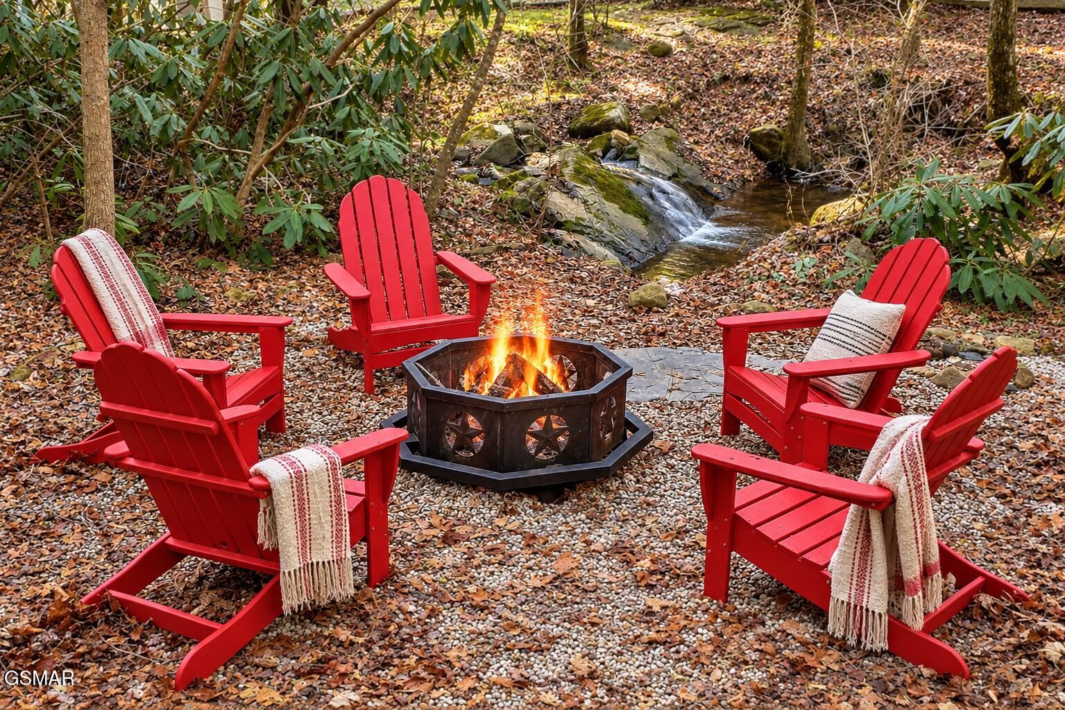 Red Adirondack chairs surround a fire pit beside a small creek in the woods.