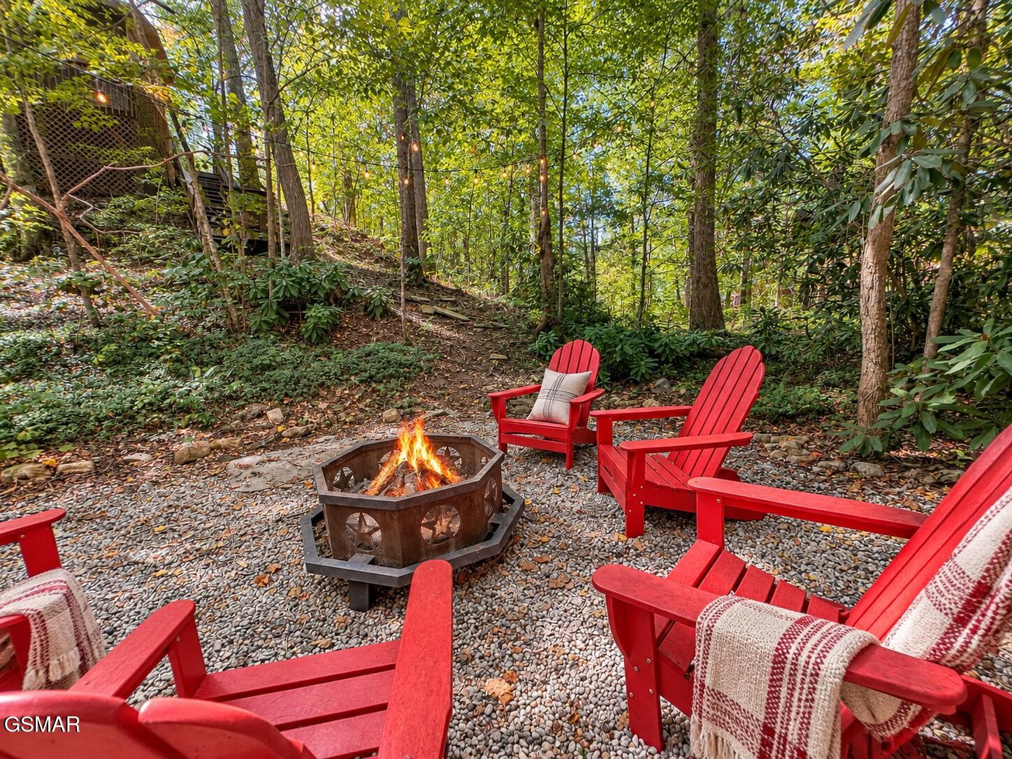 Woodland fire pit seating area with red Adirondack chairs and string lights among the trees.