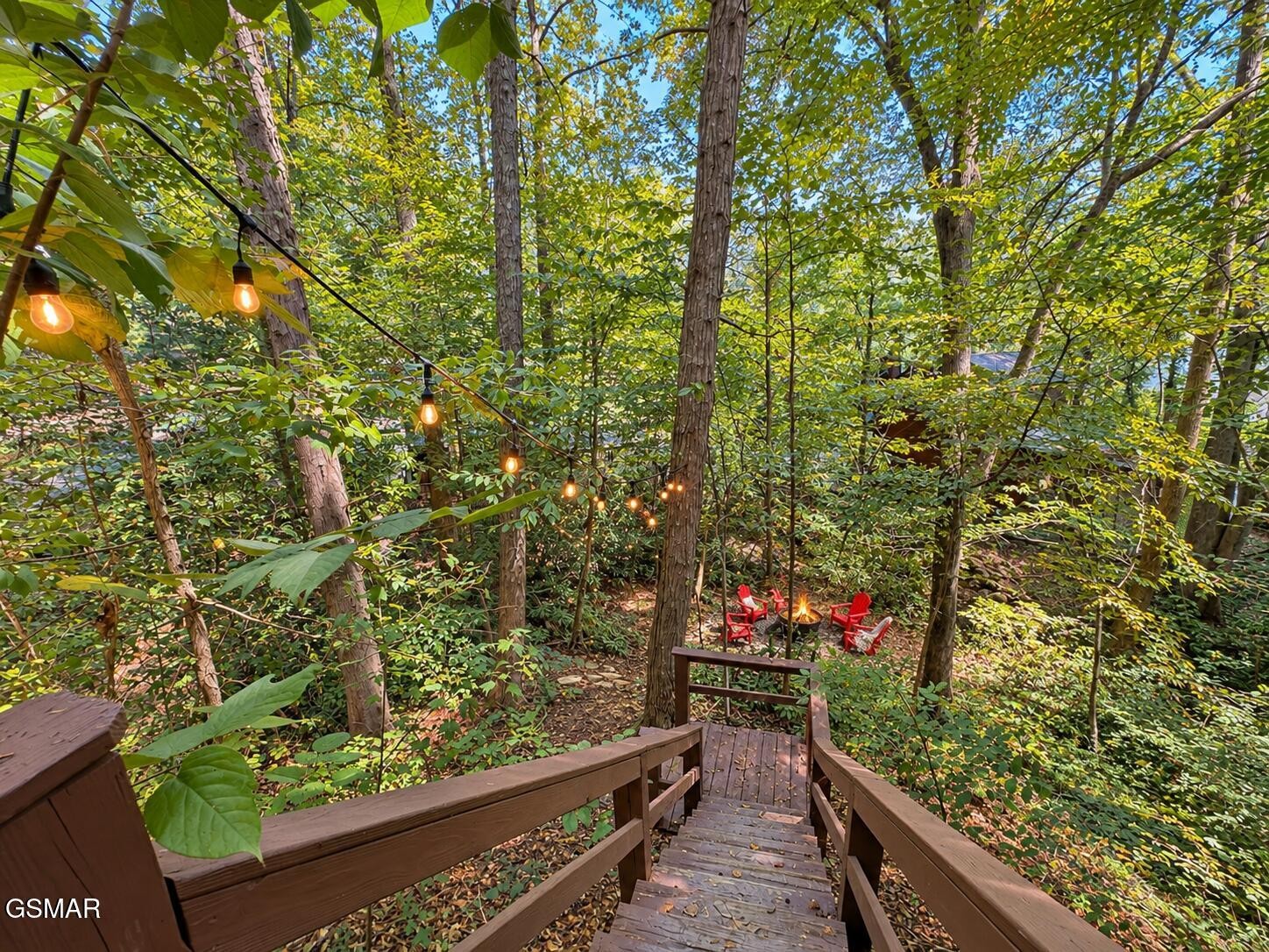 Wooden stairs descend through a wooded yard to red chairs and a fire pit beneath string lights.