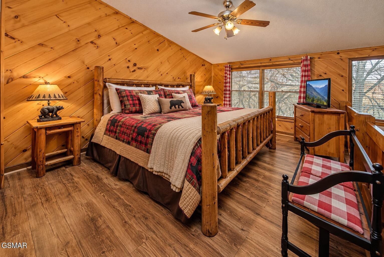 Cabin bedroom with a log-frame bed, red plaid bedding, pine walls, and chairs by the windows.