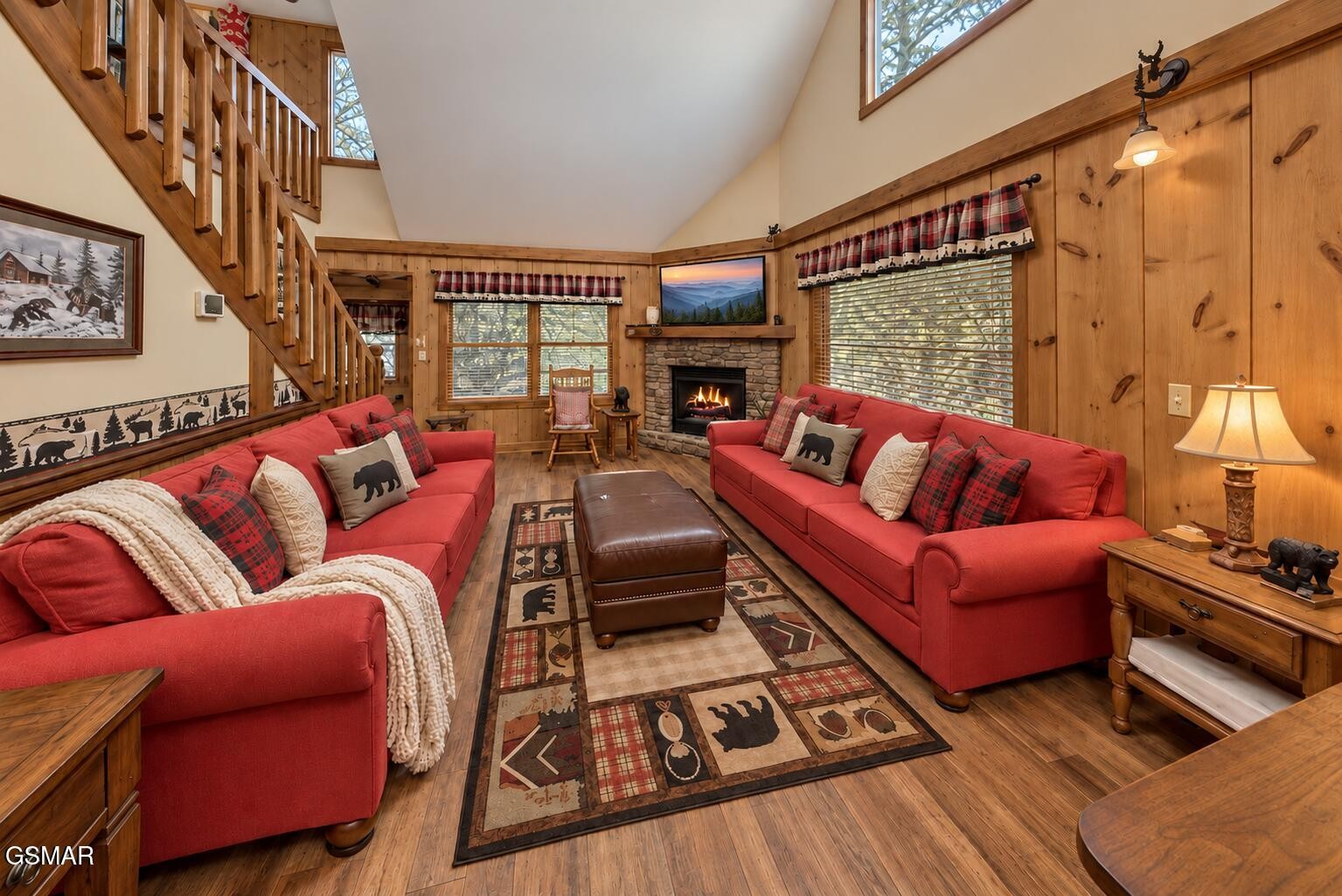 Cabin living room with red sofas, a stone fireplace, and a wooden staircase.