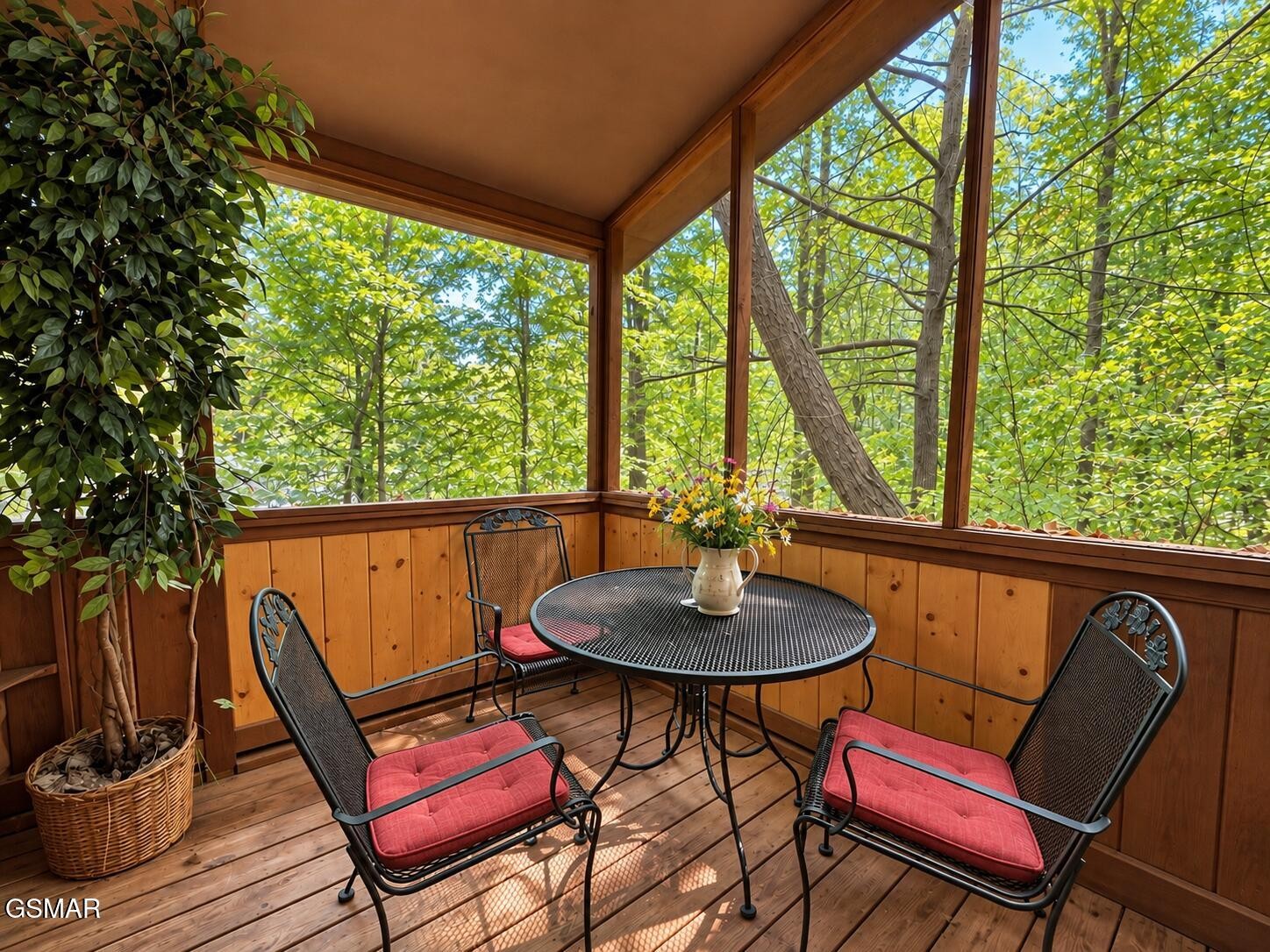 Screened porch with a round patio table, red-cushioned chairs, and leafy wooded views.