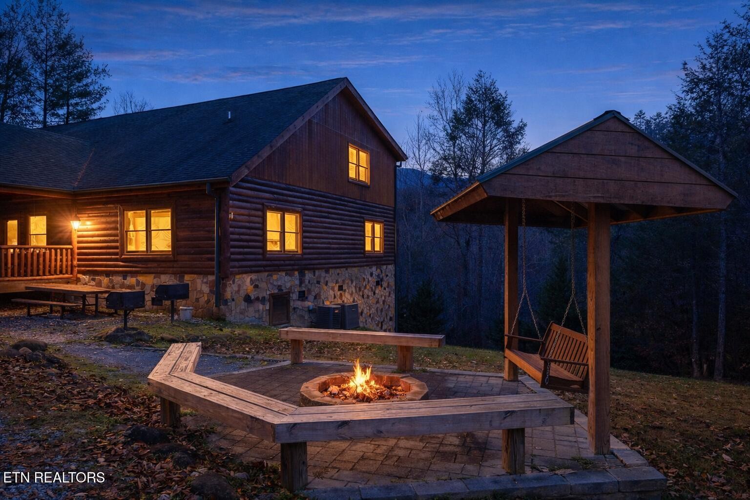 Log cabin at dusk beside a fire pit and covered wooden swing shelter.