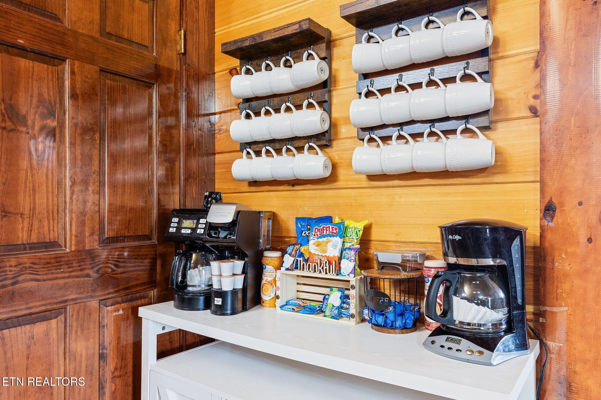 Cabin coffee station with hanging white mugs, two coffee makers, and snacks on a counter.
