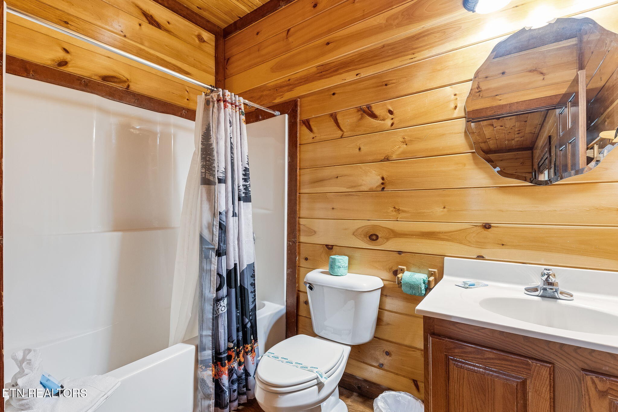 Cabin bathroom with a white tub, patterned shower curtain, toilet, and sink against wood-plank walls.