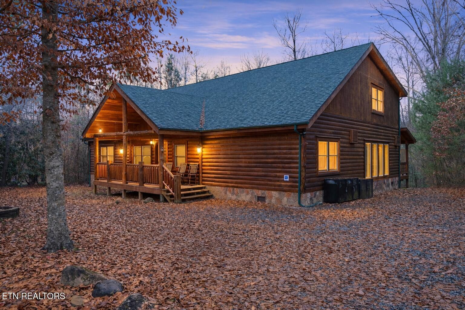 Log cabin at dusk with a covered porch, warm glowing windows, and autumn leaves carpeting the wooded lot.