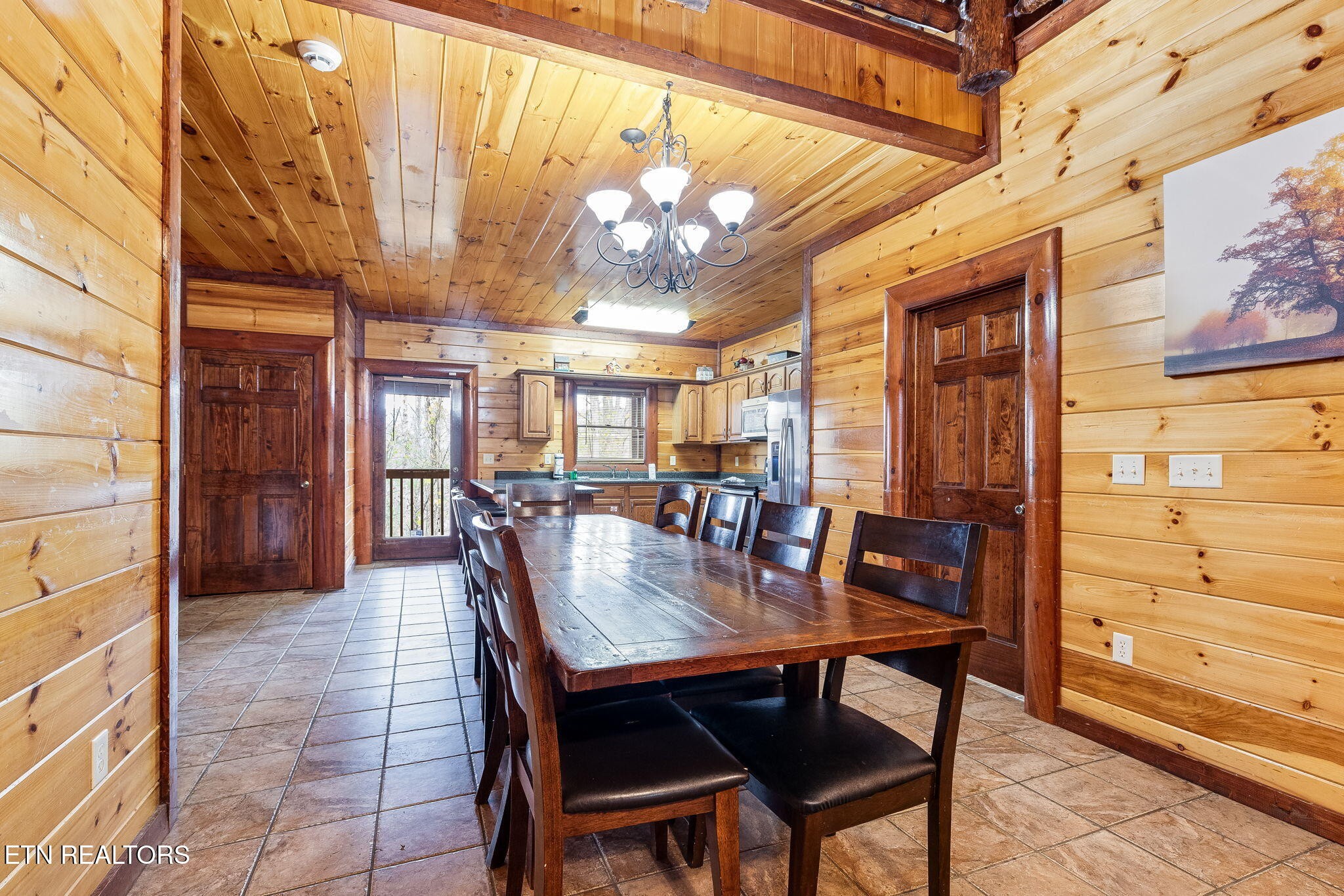 Cabin dining area with a long wooden table, exposed beams, and an adjoining kitchen.