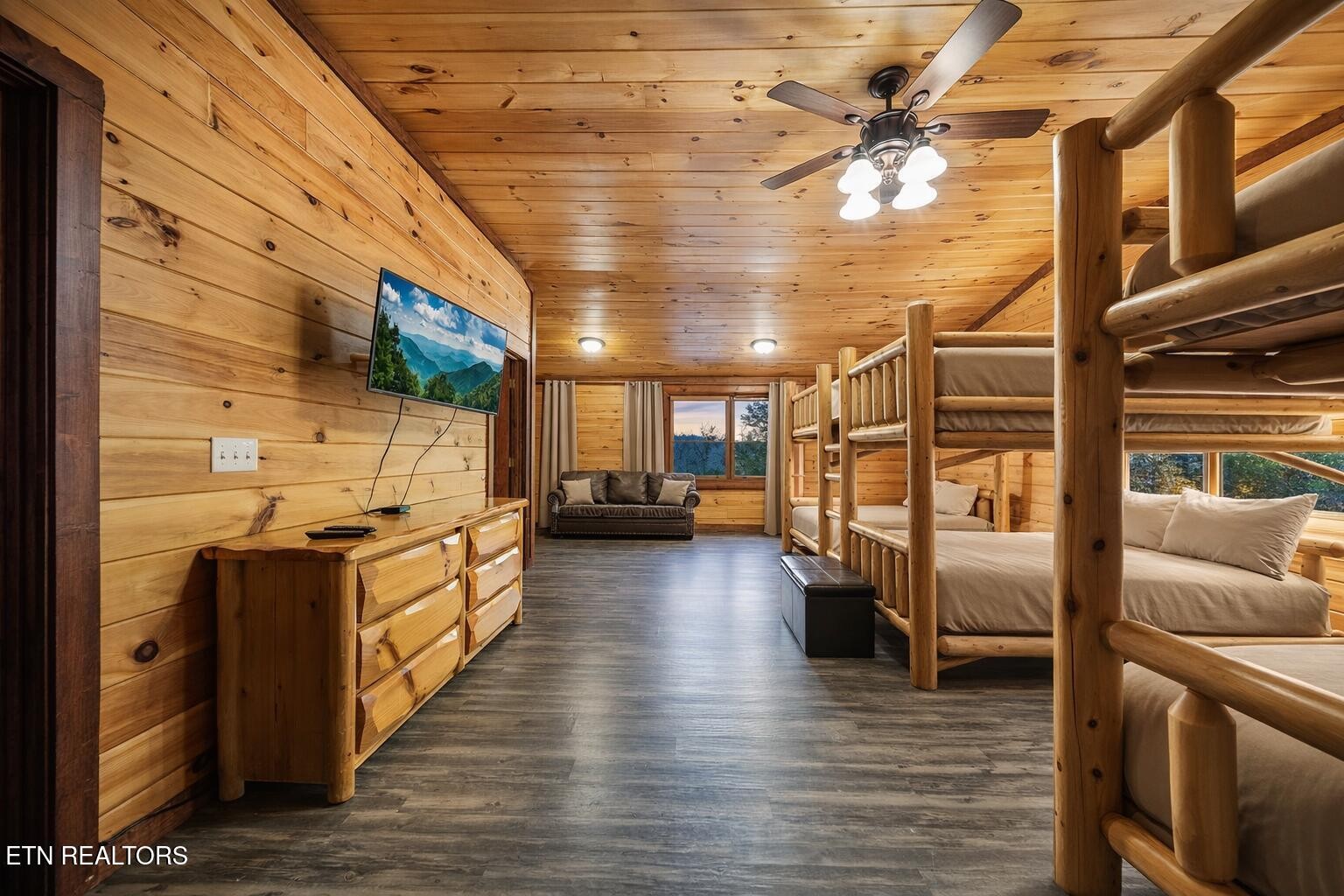 Cabin bedroom with rustic log bunk beds, wood-paneled walls, and a sofa beneath the windows.