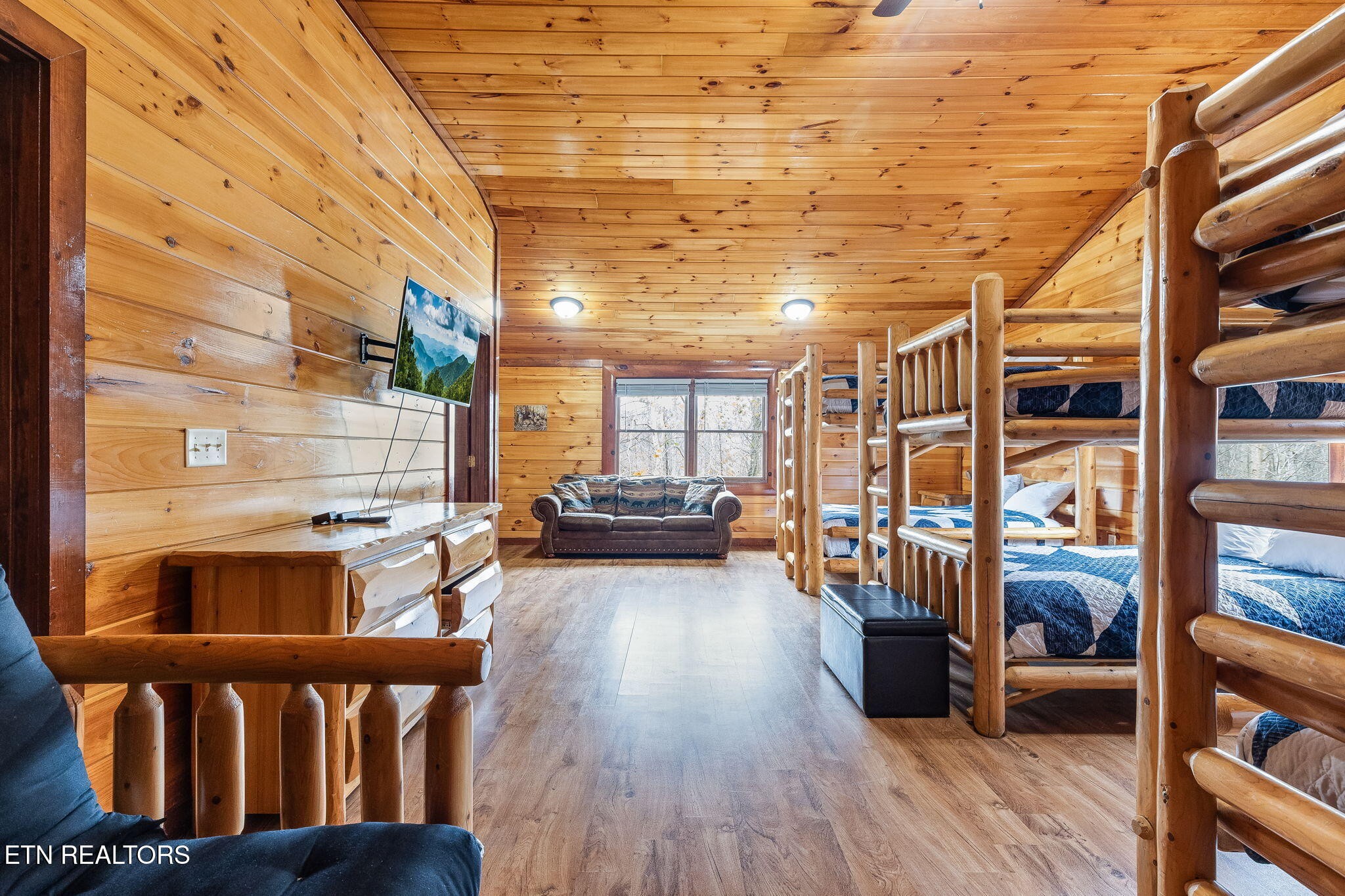 Cabin bedroom with rustic log bunk beds, blue-and-white bedding, and a sofa beneath the window.