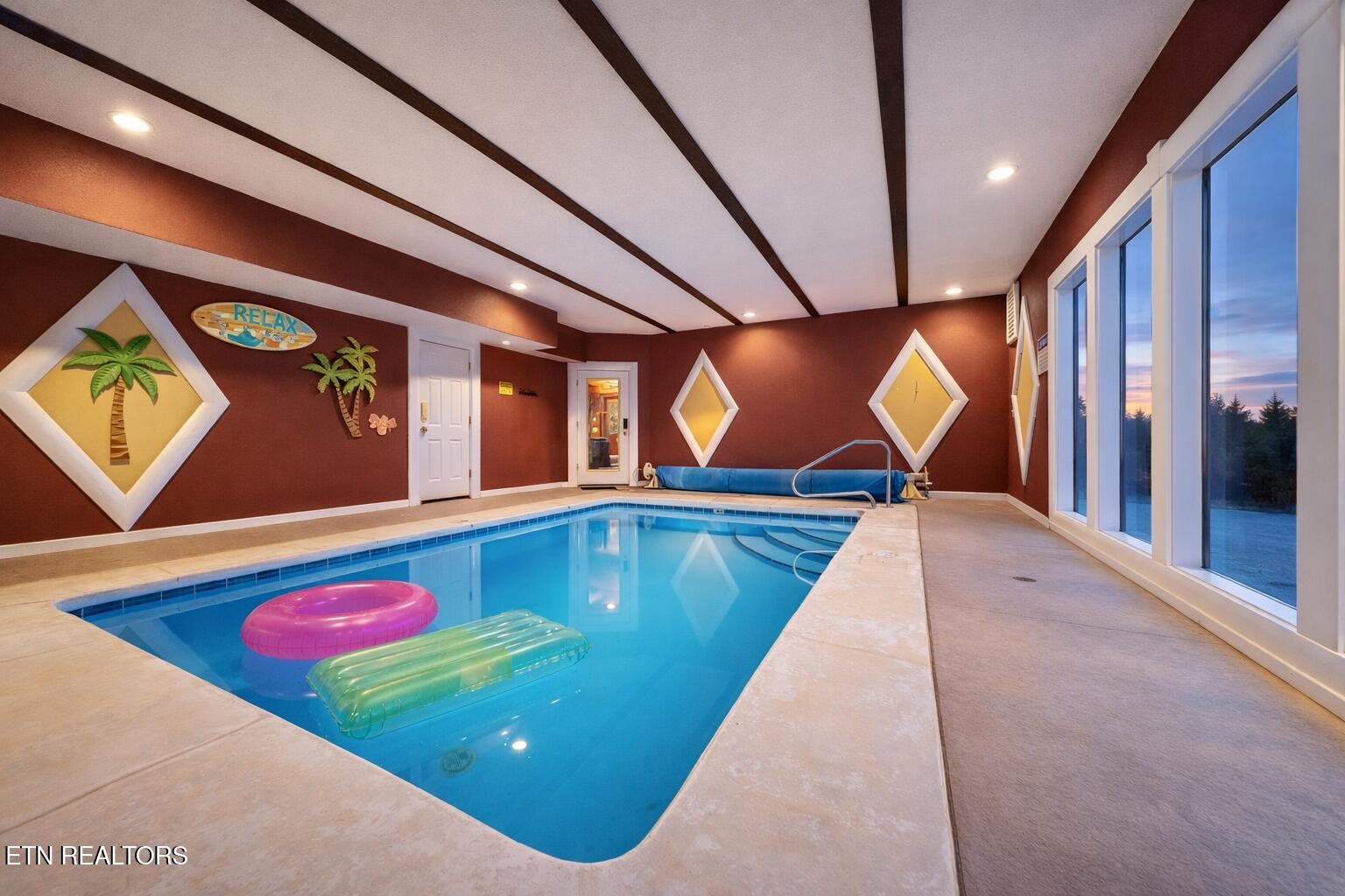 Indoor swimming pool with colorful floats, tropical wall decor, and tall windows.