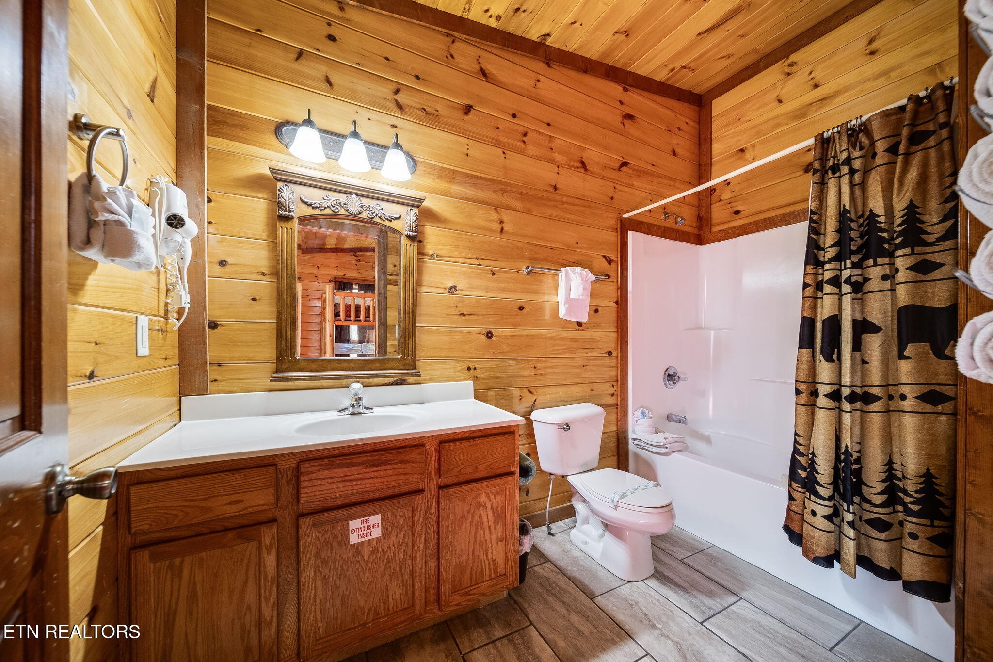 Cabin bathroom with warm wood-paneled walls, a wooden vanity, and a forest-patterned shower curtain.