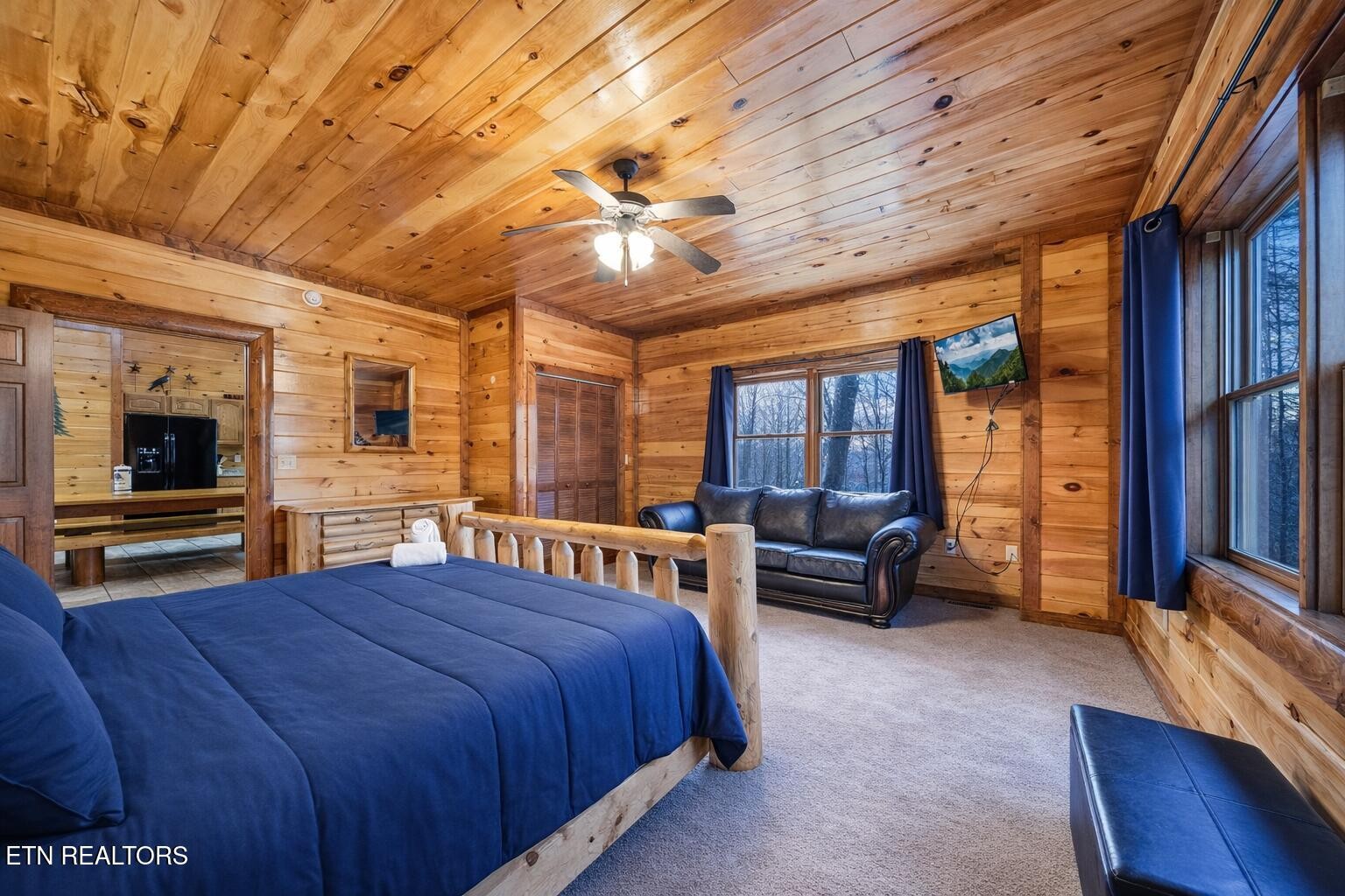 Cabin bedroom with a large blue bed, wood-paneled walls, and a leather loveseat beside wooded windows.