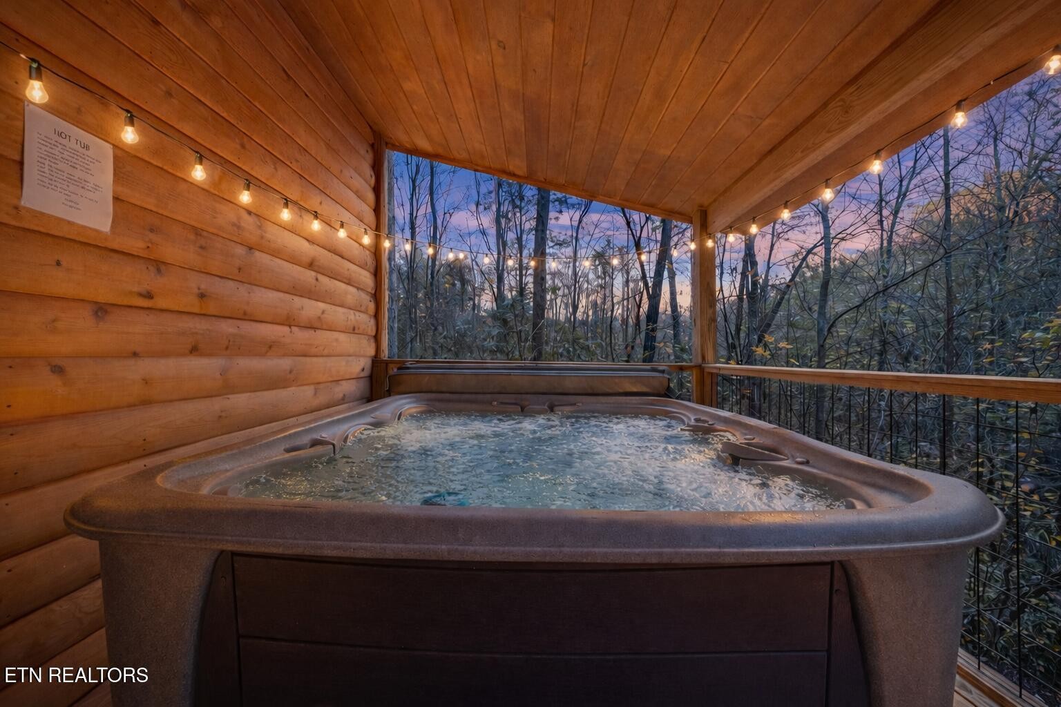 Hot tub on a covered wooden porch overlooking leafless woods at dusk.