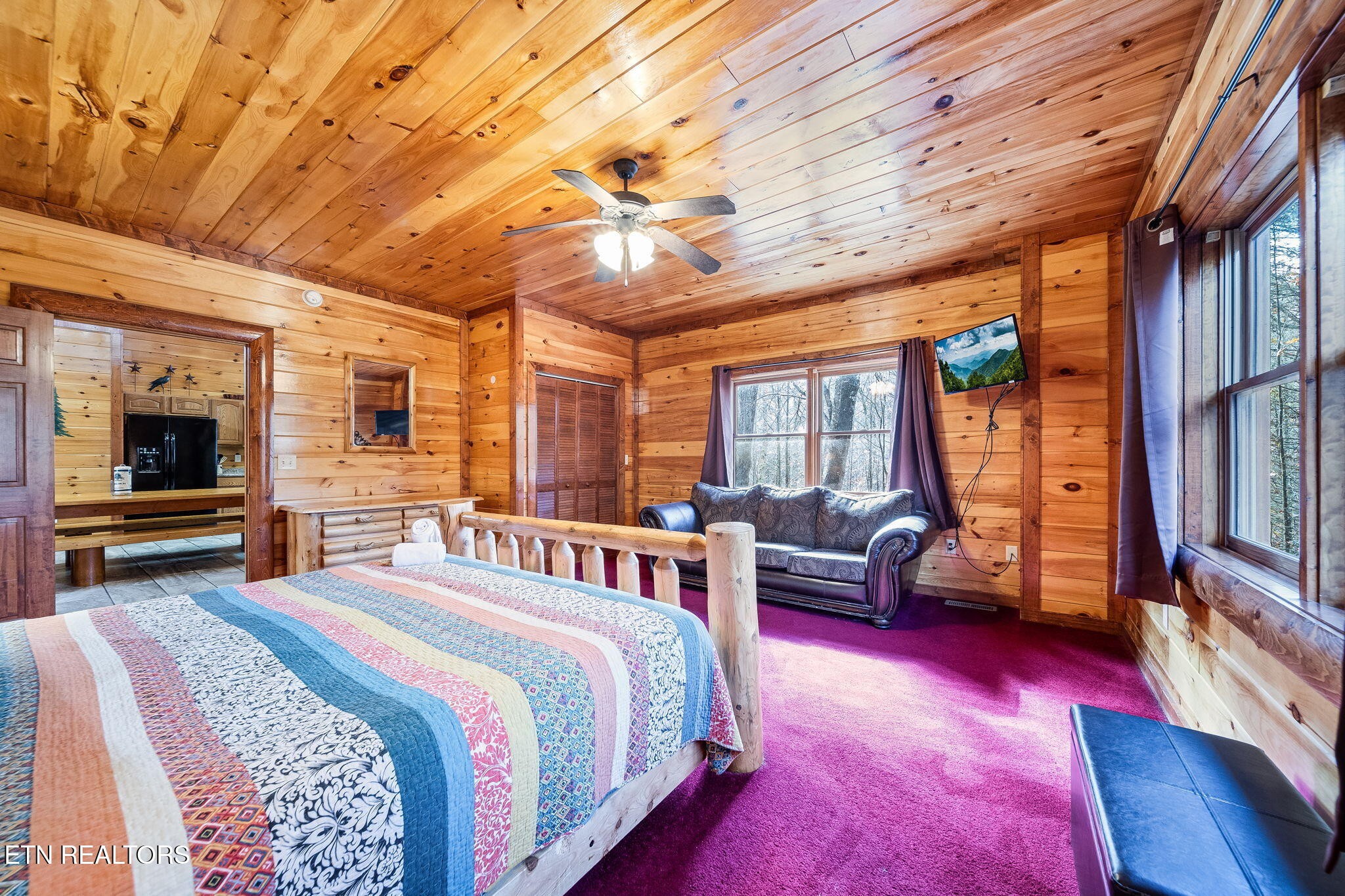 Cabin bedroom with a log-style bed, colorful quilt, wood-paneled walls, and a loveseat by the window.