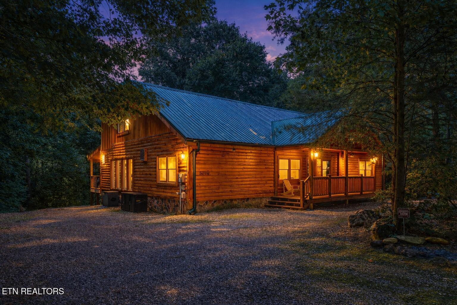 Wooded log cabin at dusk with a blue metal roof and warmly lit covered porch.