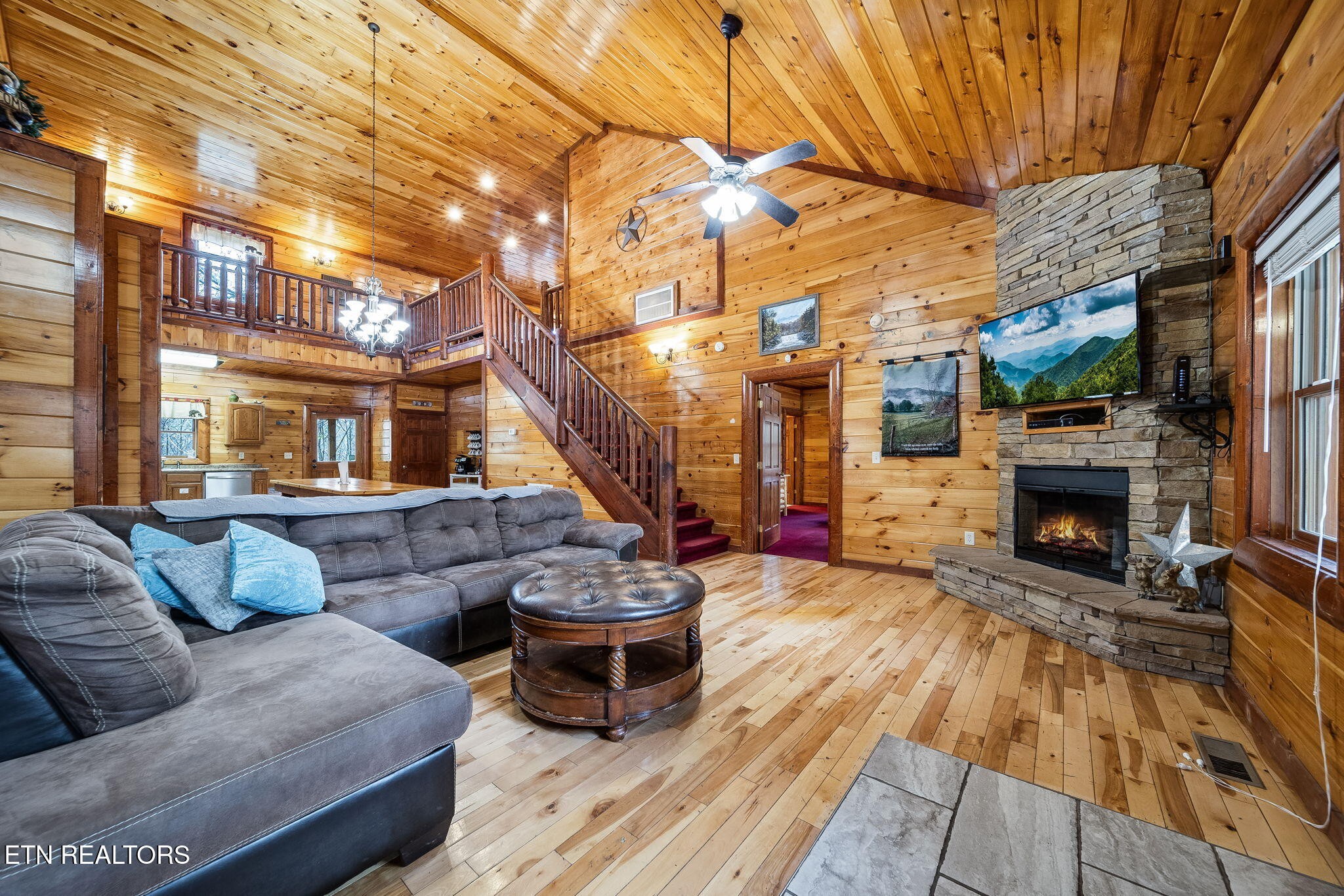 Cabin living room with a vaulted wood ceiling, loft staircase, gray sectional, and stone fireplace.