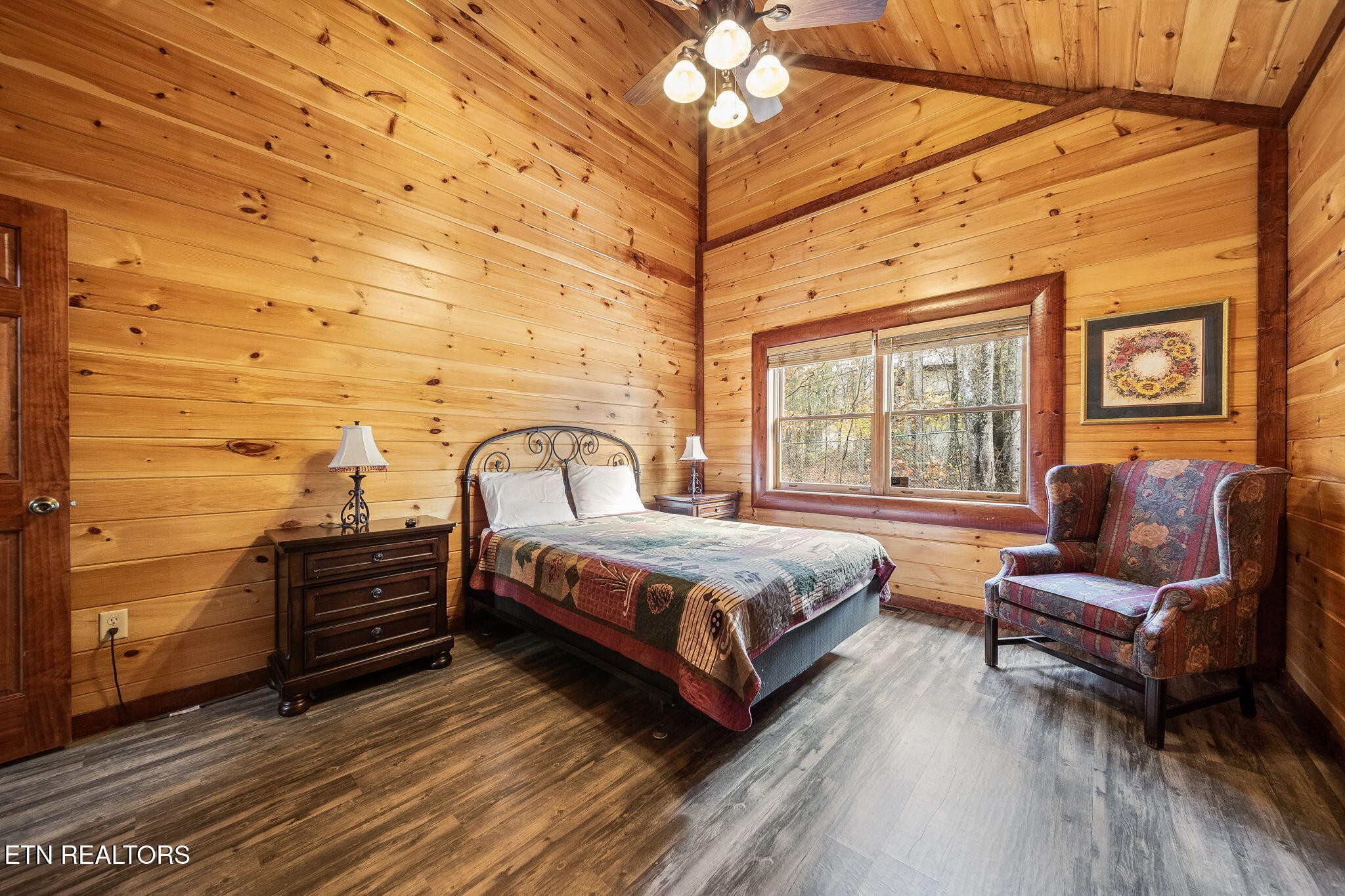 Cabin bedroom with wood-paneled walls, a patterned bedspread, and an armchair beside a large window.