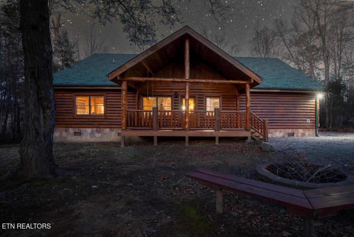 Log cabin with a covered porch and glowing windows beside a gravel driveway and fire pit.