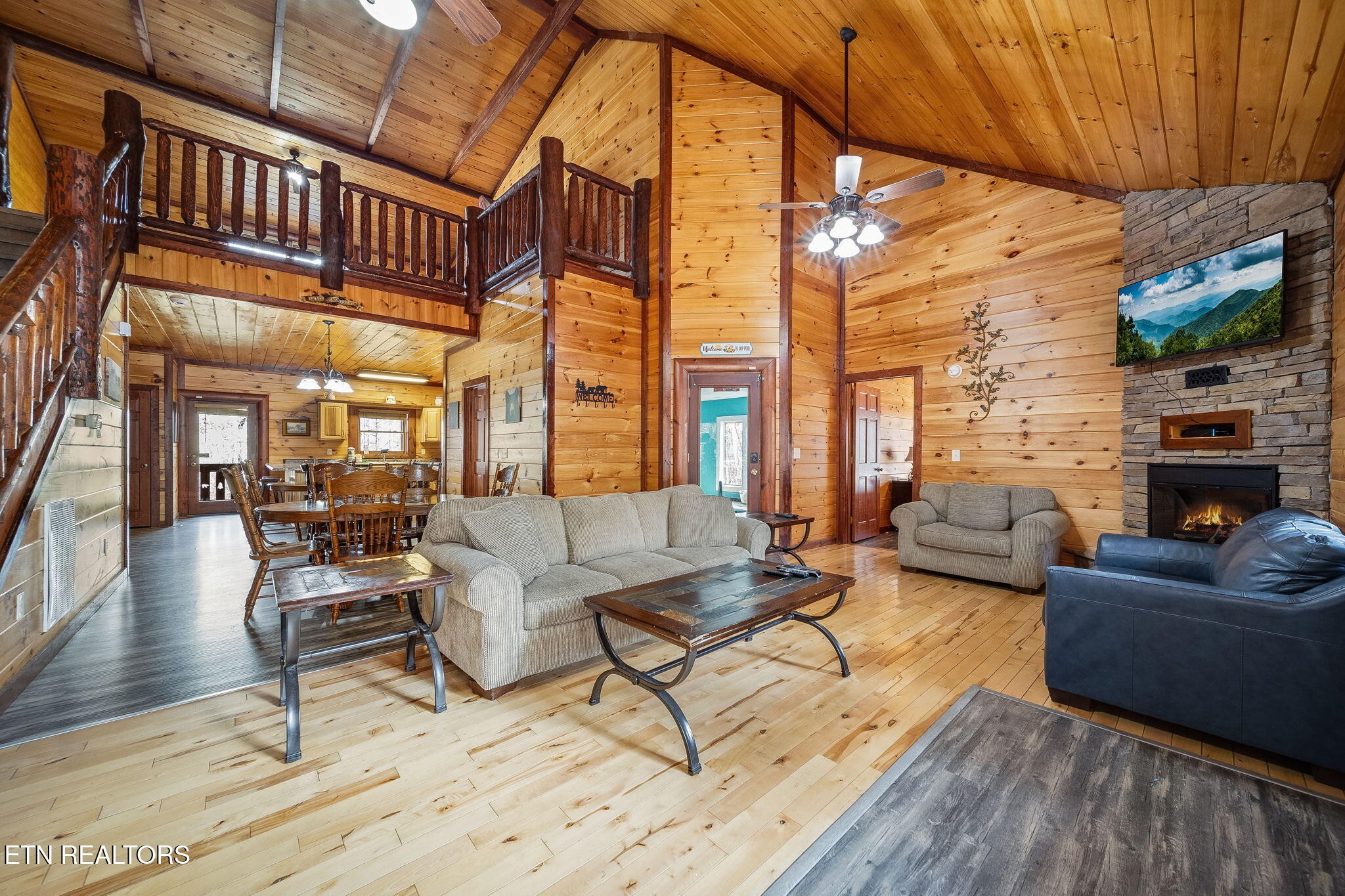 Cabin living room with a vaulted wood ceiling, loft railing, dining area, and stone fireplace.