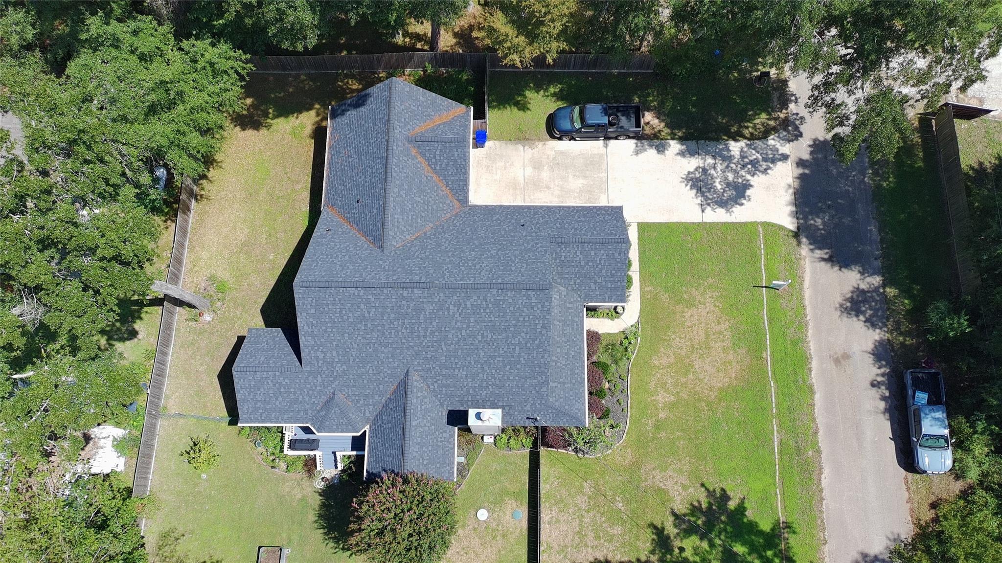 Aerial view of a home with a broad gray roof, driveway, lawn, and mature trees.