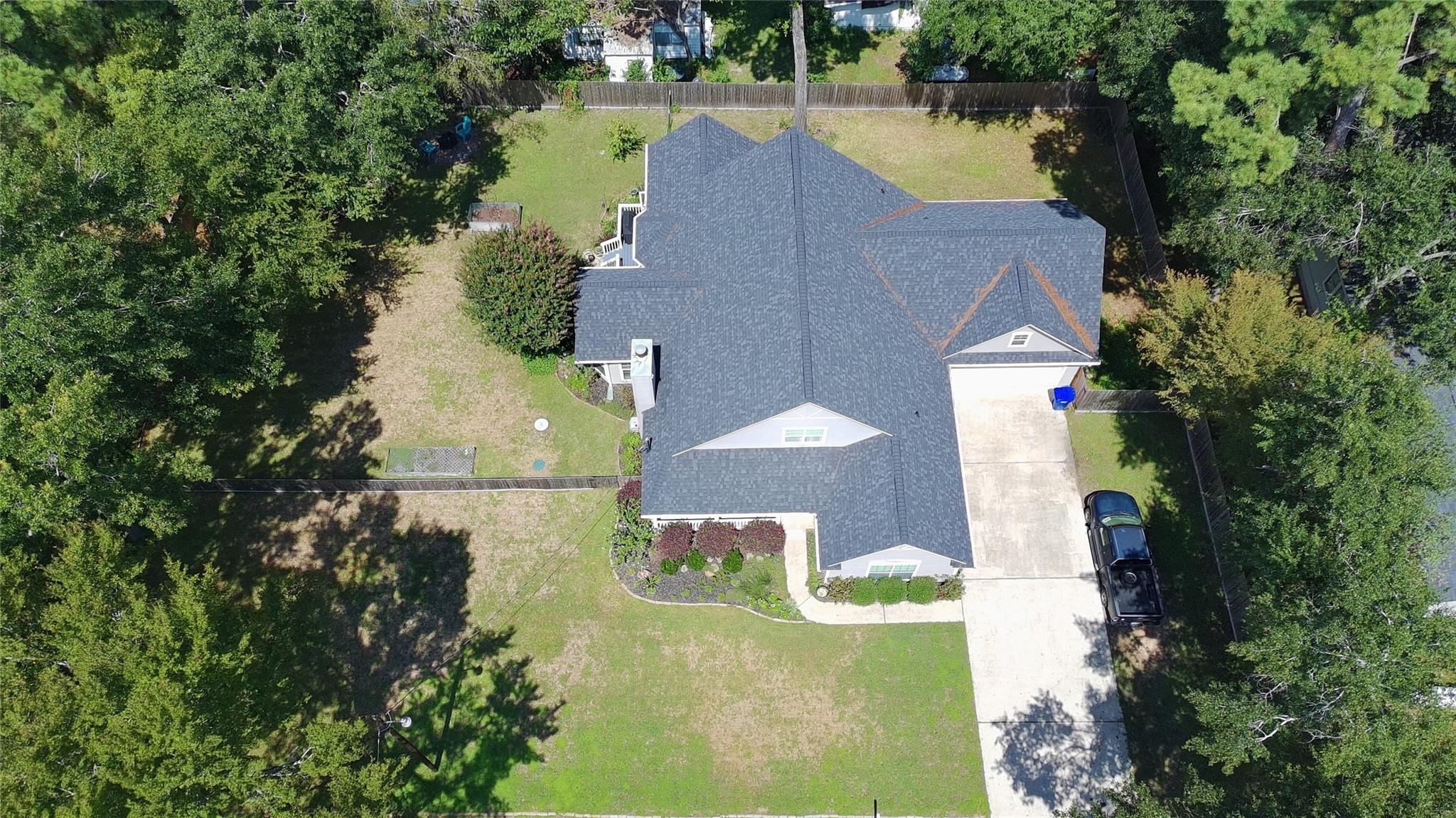 Aerial view of a home with a gray roof, fenced yard, mature trees, and a long driveway.
