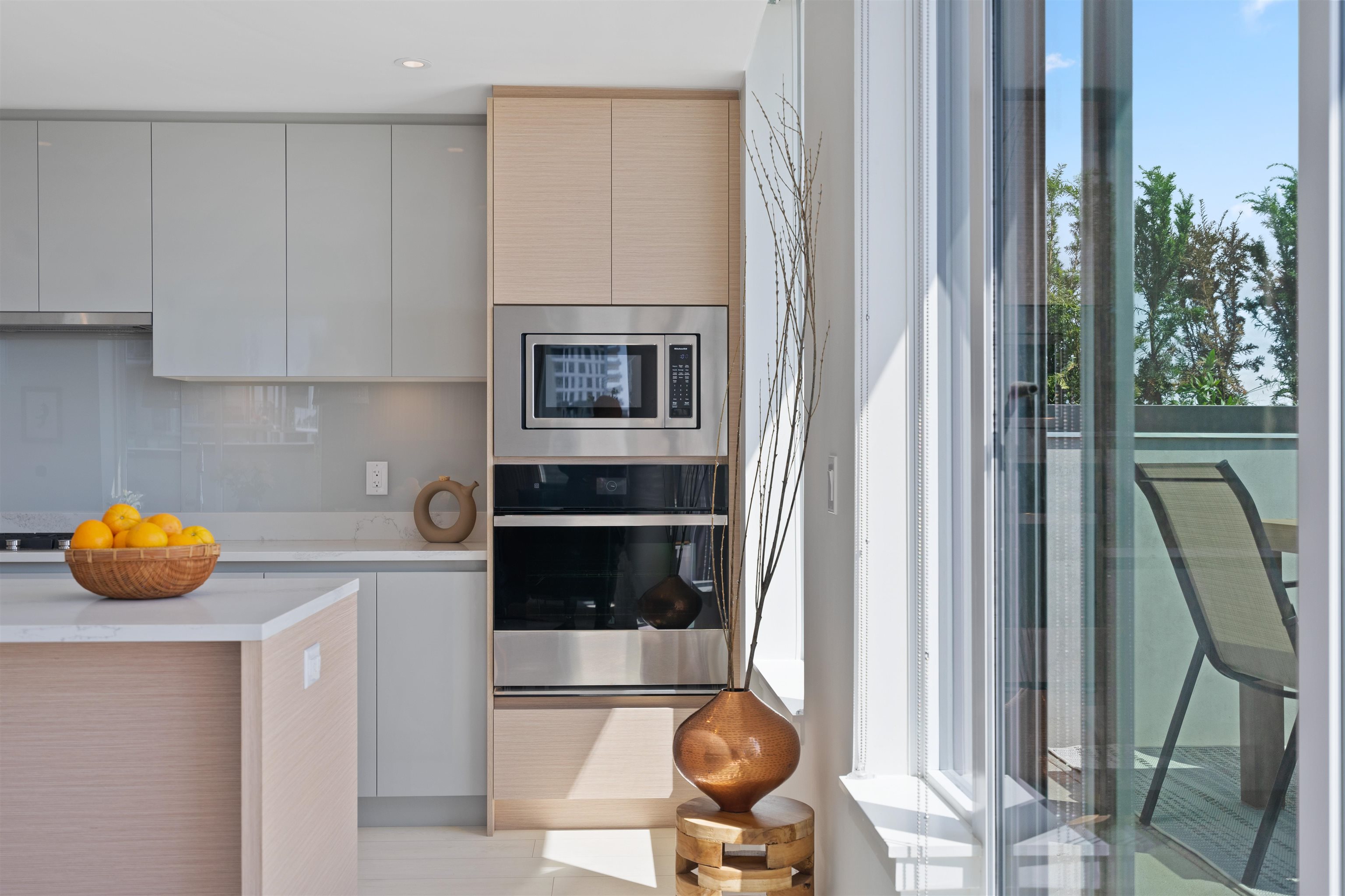 Modern kitchen with pale wood cabinetry, built-in appliances, and windows overlooking a balcony.