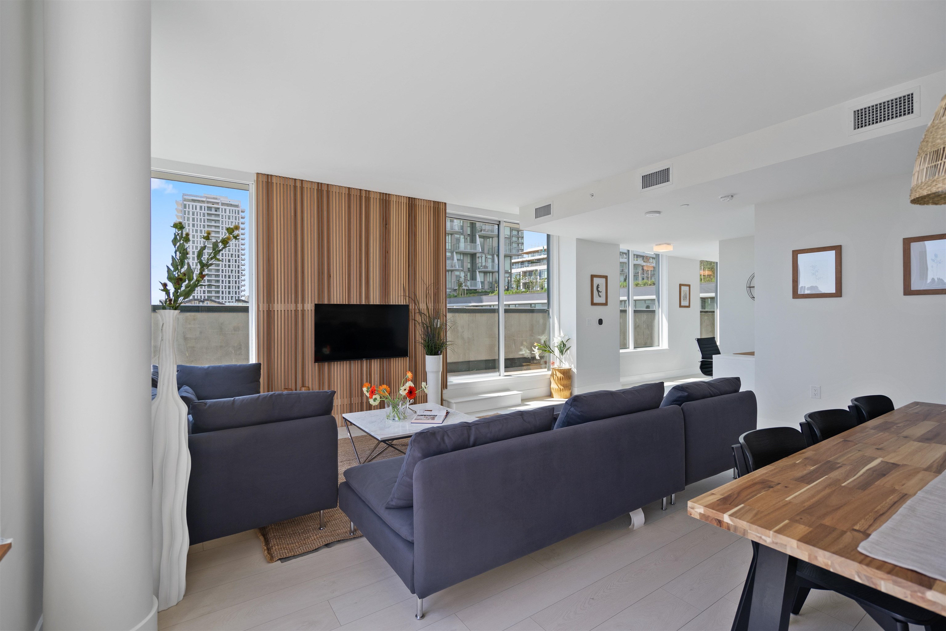 Open-plan living and dining area with dark sofas, a wood-slat TV wall, and large windows.
