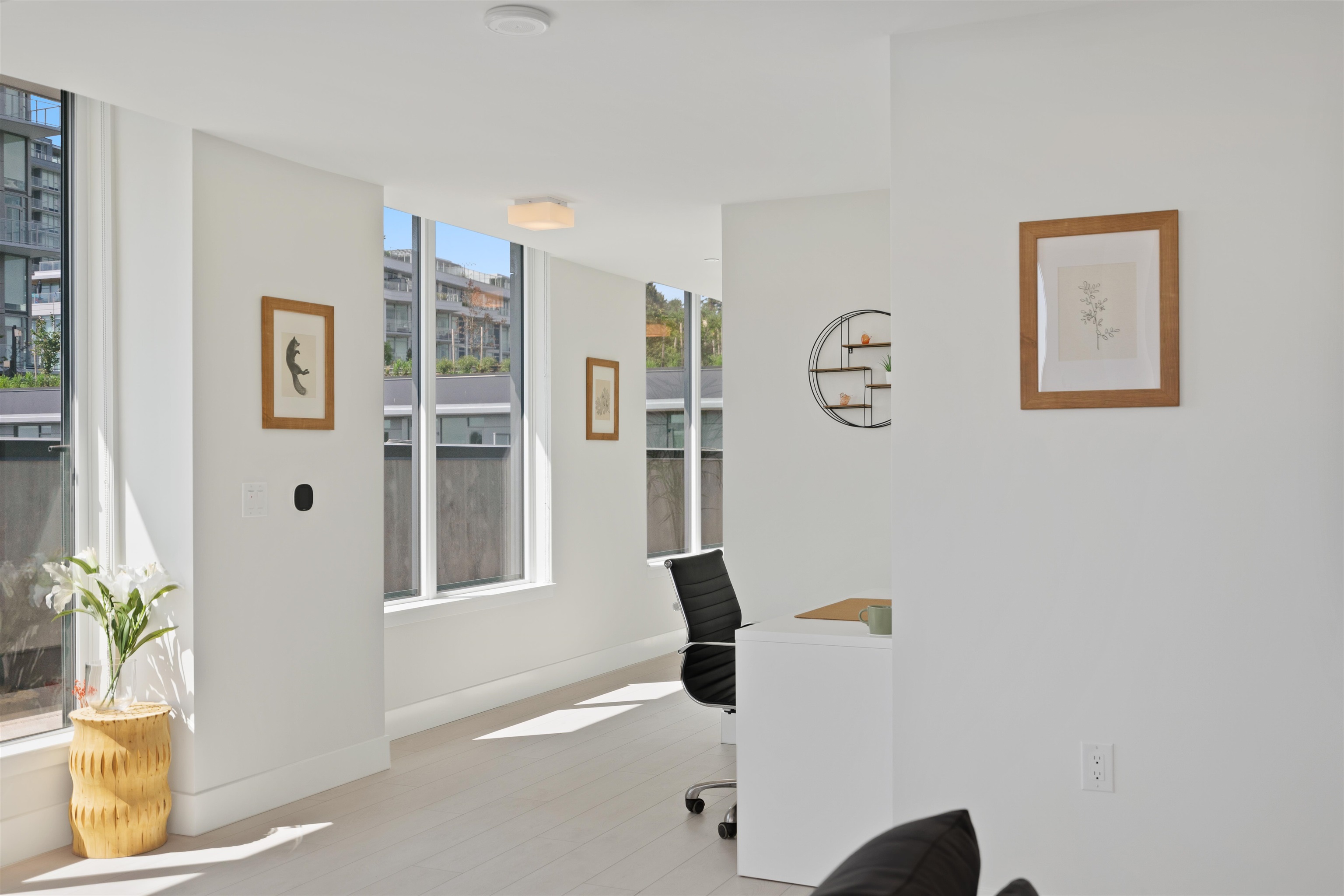 Bright home office with a white desk, black chair, and large windows overlooking neighboring buildings.
