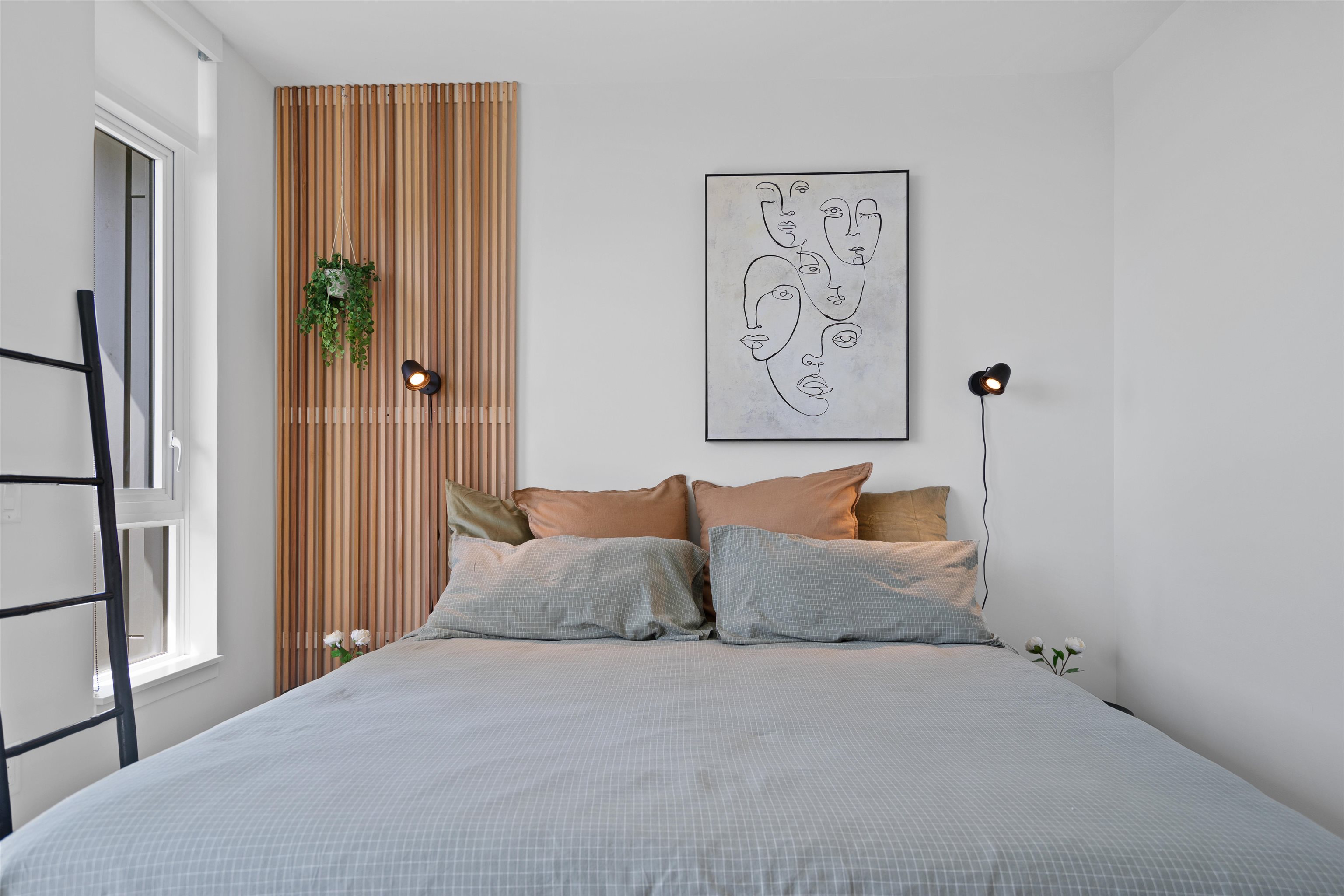 Staged bedroom with a large gray-checked bed, wood-slat feature wall, hanging plant, and framed line-art artwork.