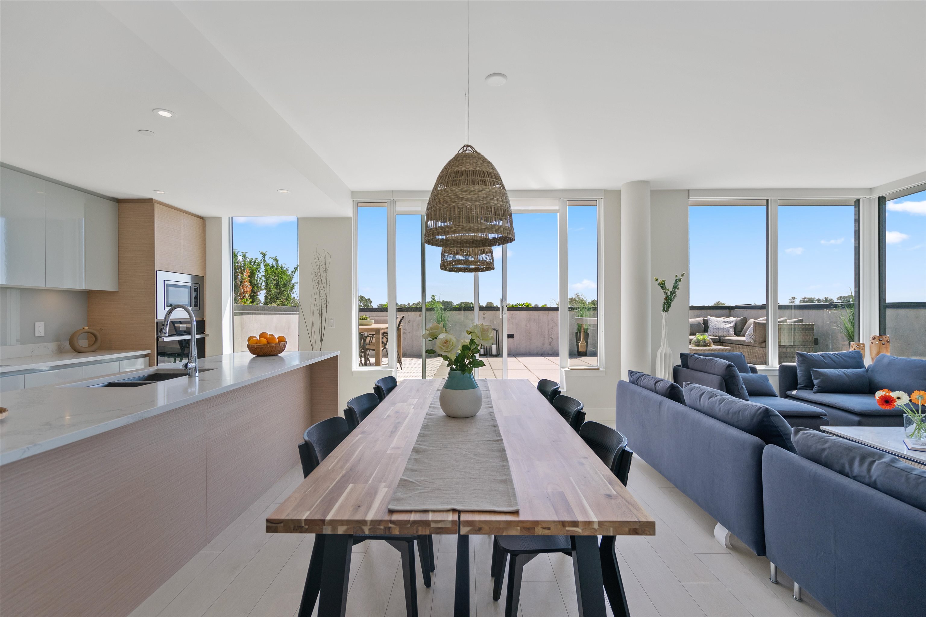 Open-plan dining and living room with a long wood table, blue sofas, and windows onto a terrace.