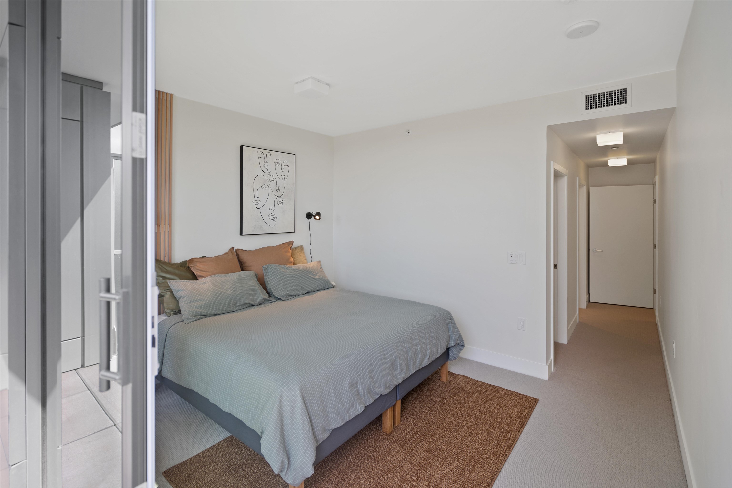 Bedroom with gray-blue bedding, layered pillows, framed line art, and a hallway beside the bed.