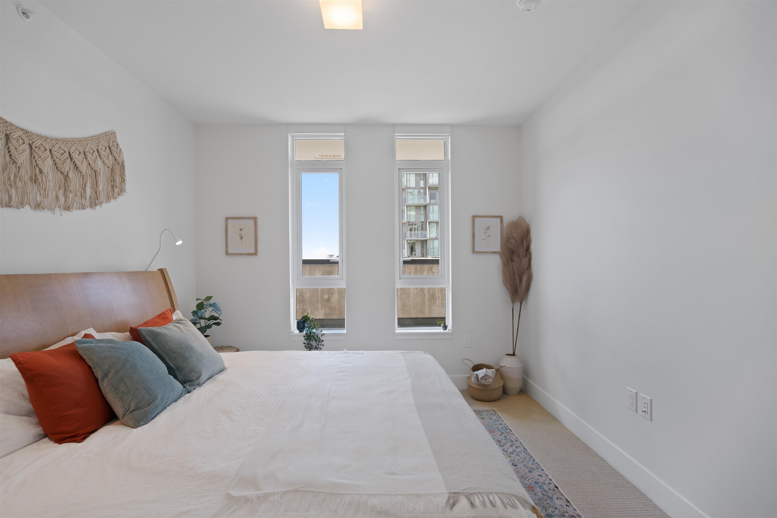 Staged bedroom with a large bed and two tall windows overlooking a neighboring building.