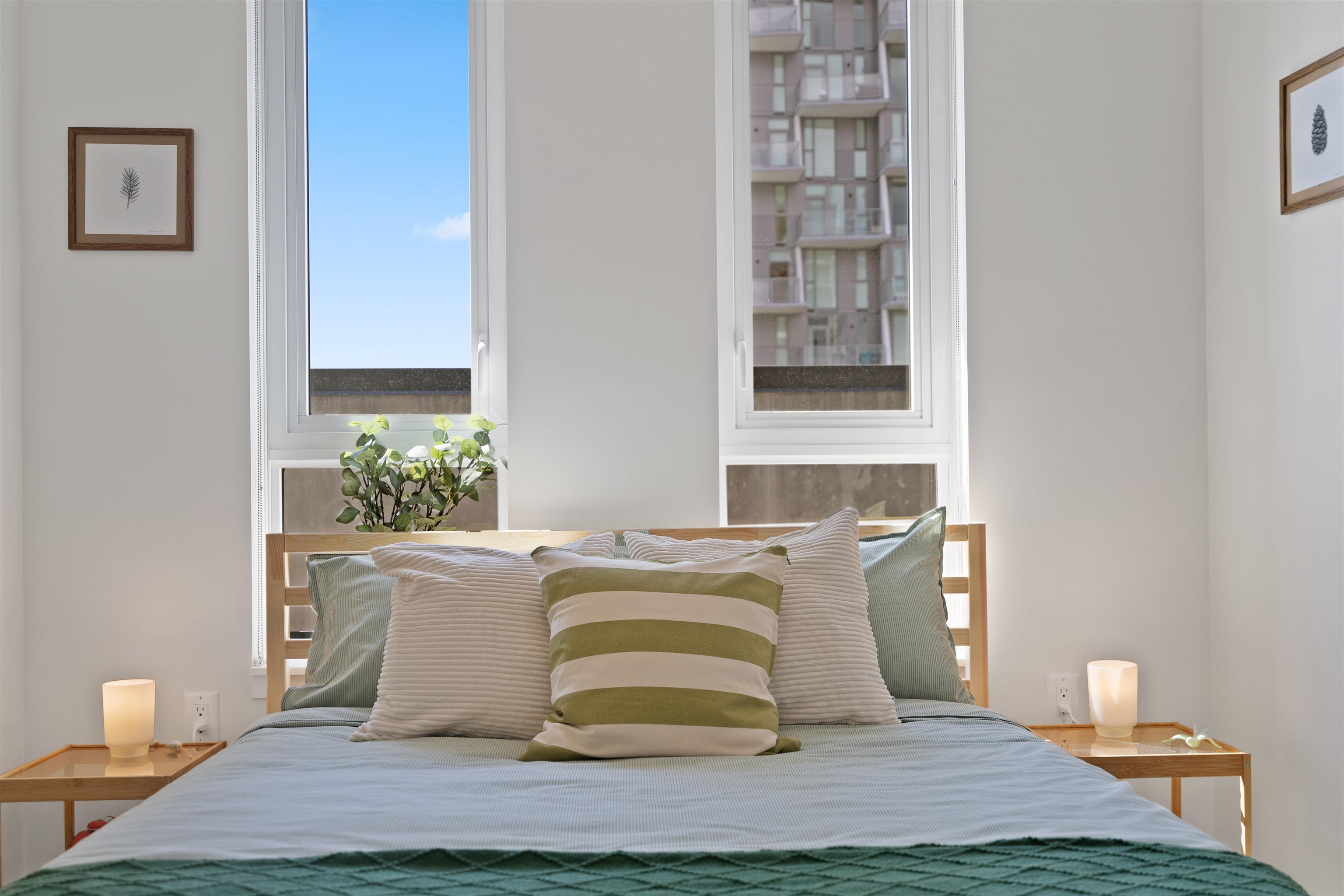 Staged bedroom with a blue-and-green bed, tall windows, and a neighboring apartment building outside.