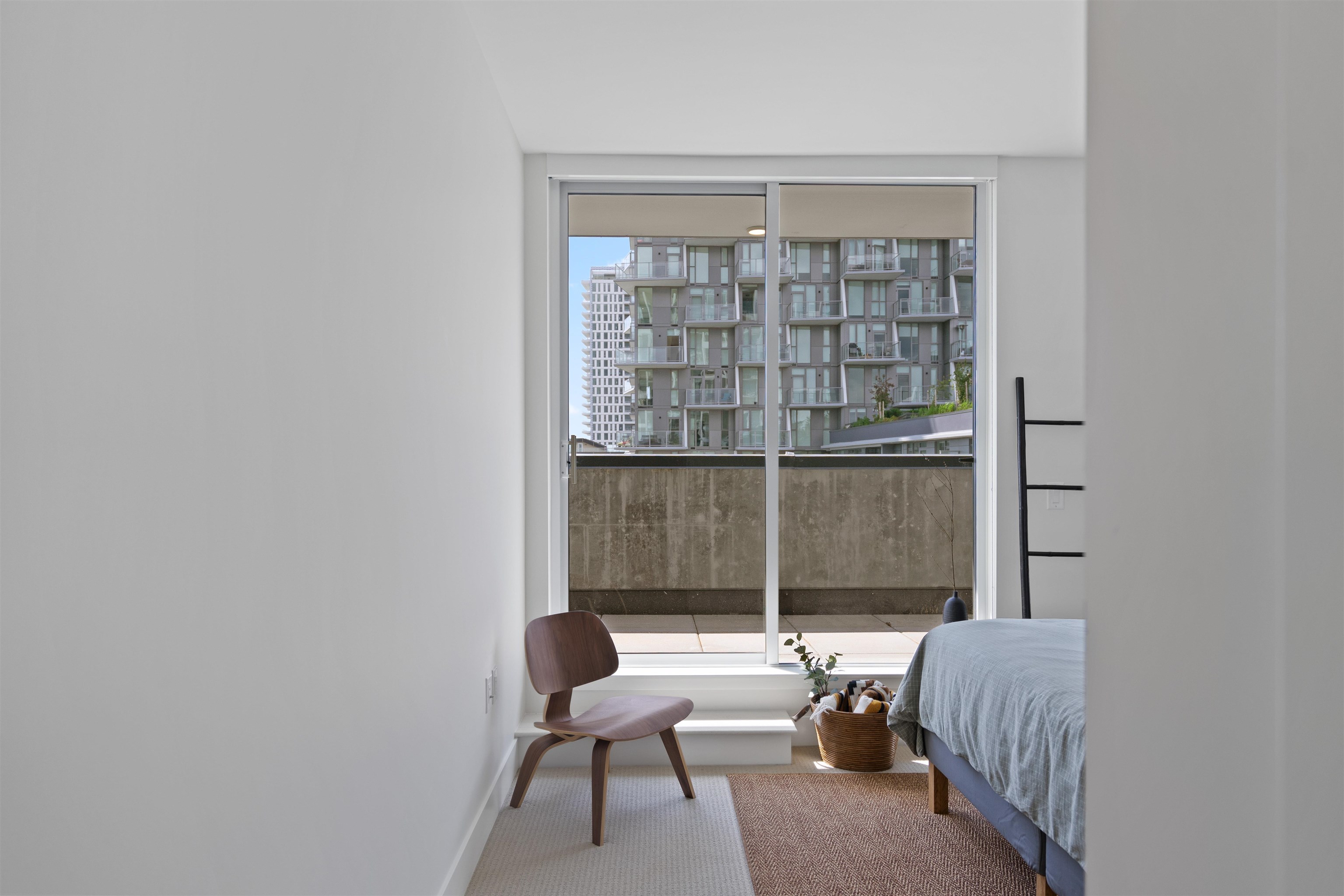 Furnished bedroom with a large window overlooking a modern apartment building and balcony.