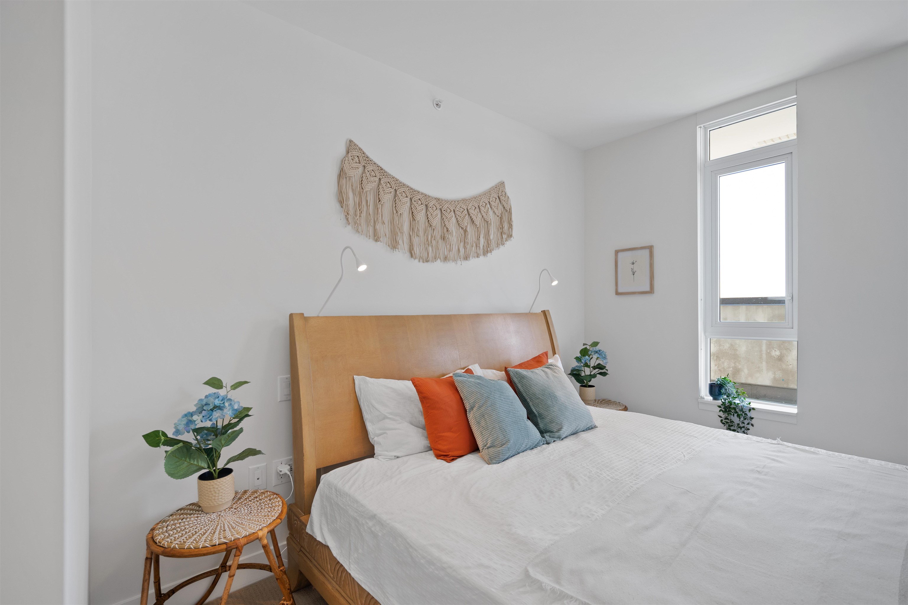 Bedroom with a light wood headboard, colorful pillows, and a tall window.
