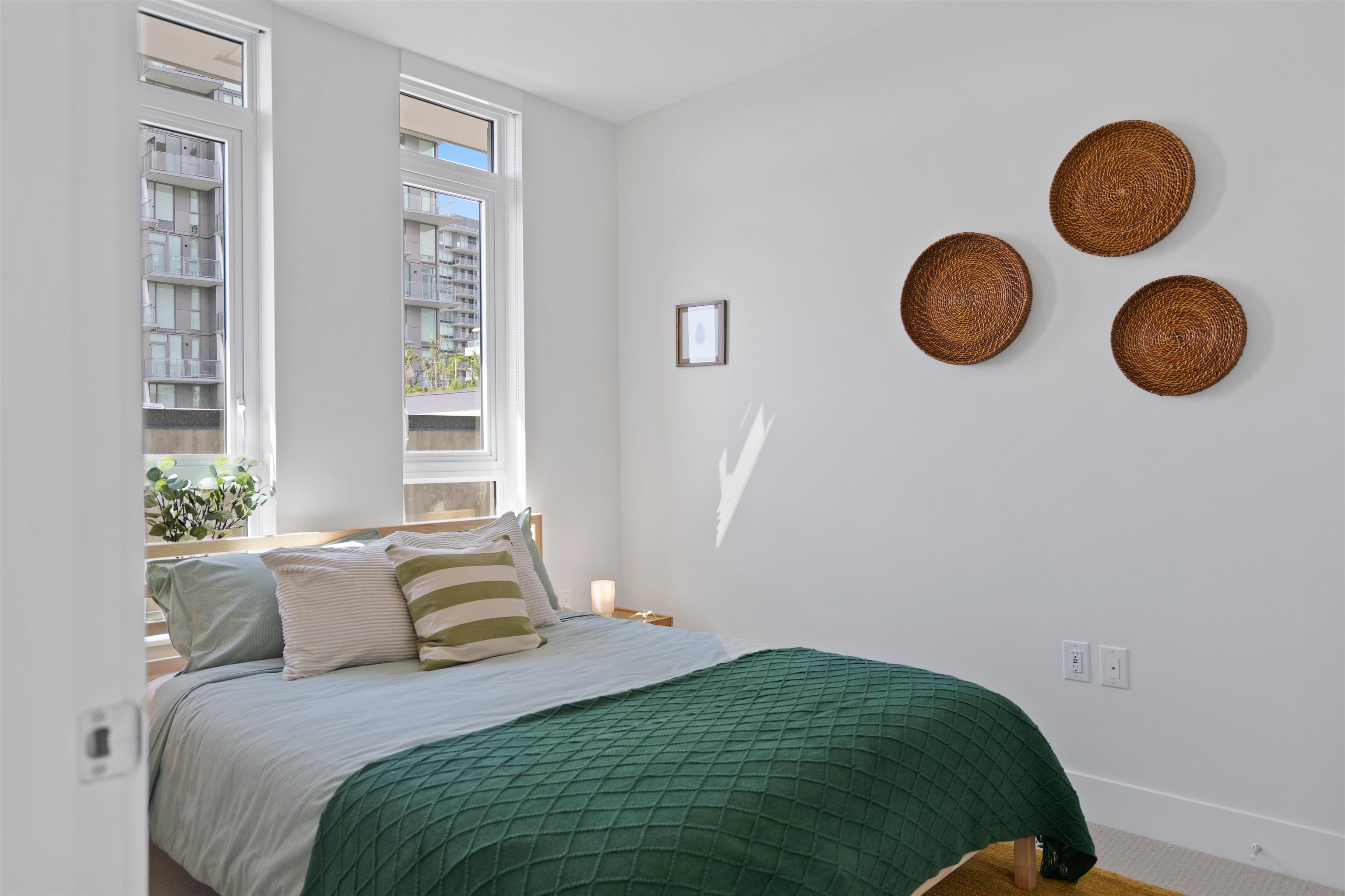 Staged bedroom with a green quilt, tall windows, and woven wall decorations.