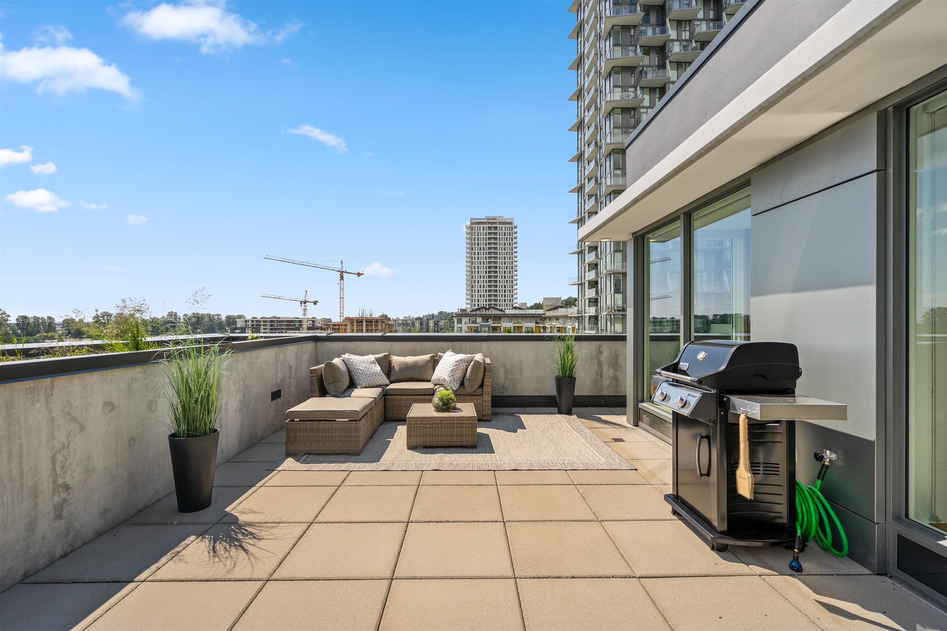 Condo terrace with wicker seating, potted plants, and a barbecue grill overlooking nearby high-rises.