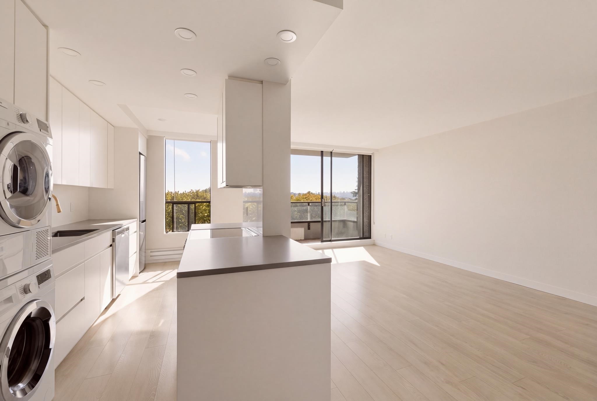 Vacant open-plan kitchen with a large island, stacked laundry appliances, and sunny balcony windows.