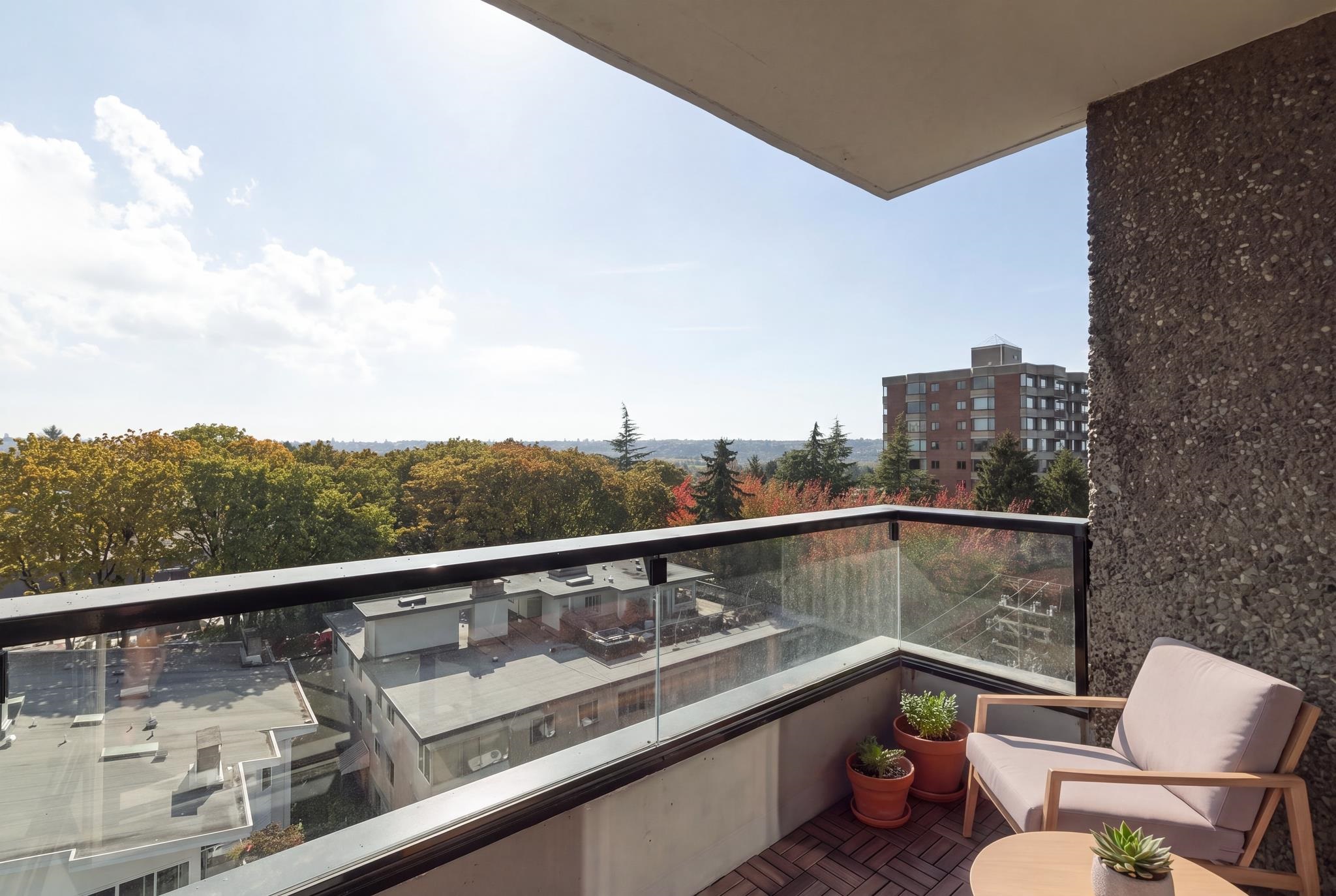 Apartment balcony with glass railings, potted plants, and an autumn treetop view.