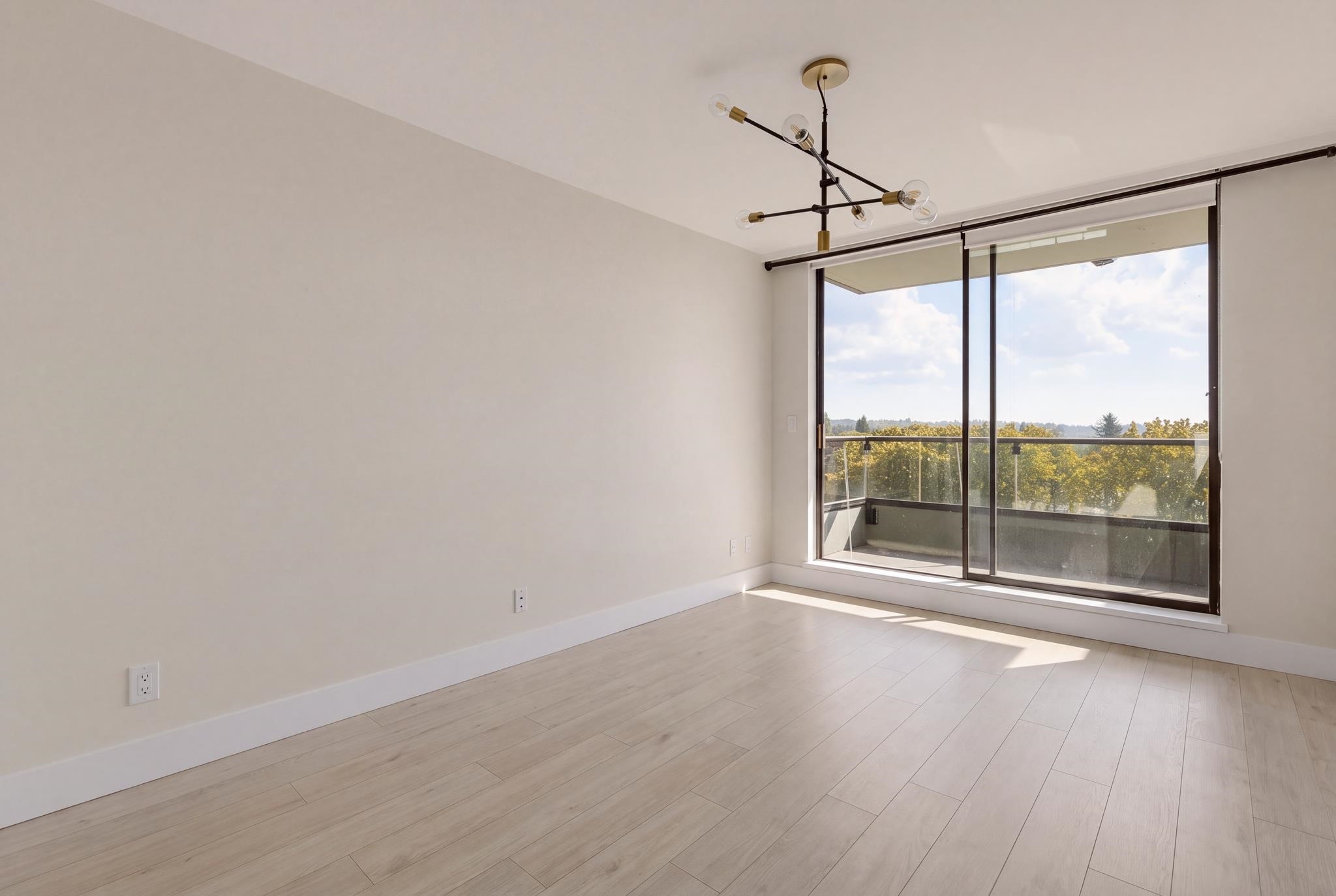 Empty living room with pale plank floors, a modern chandelier, and sliding doors to a balcony.