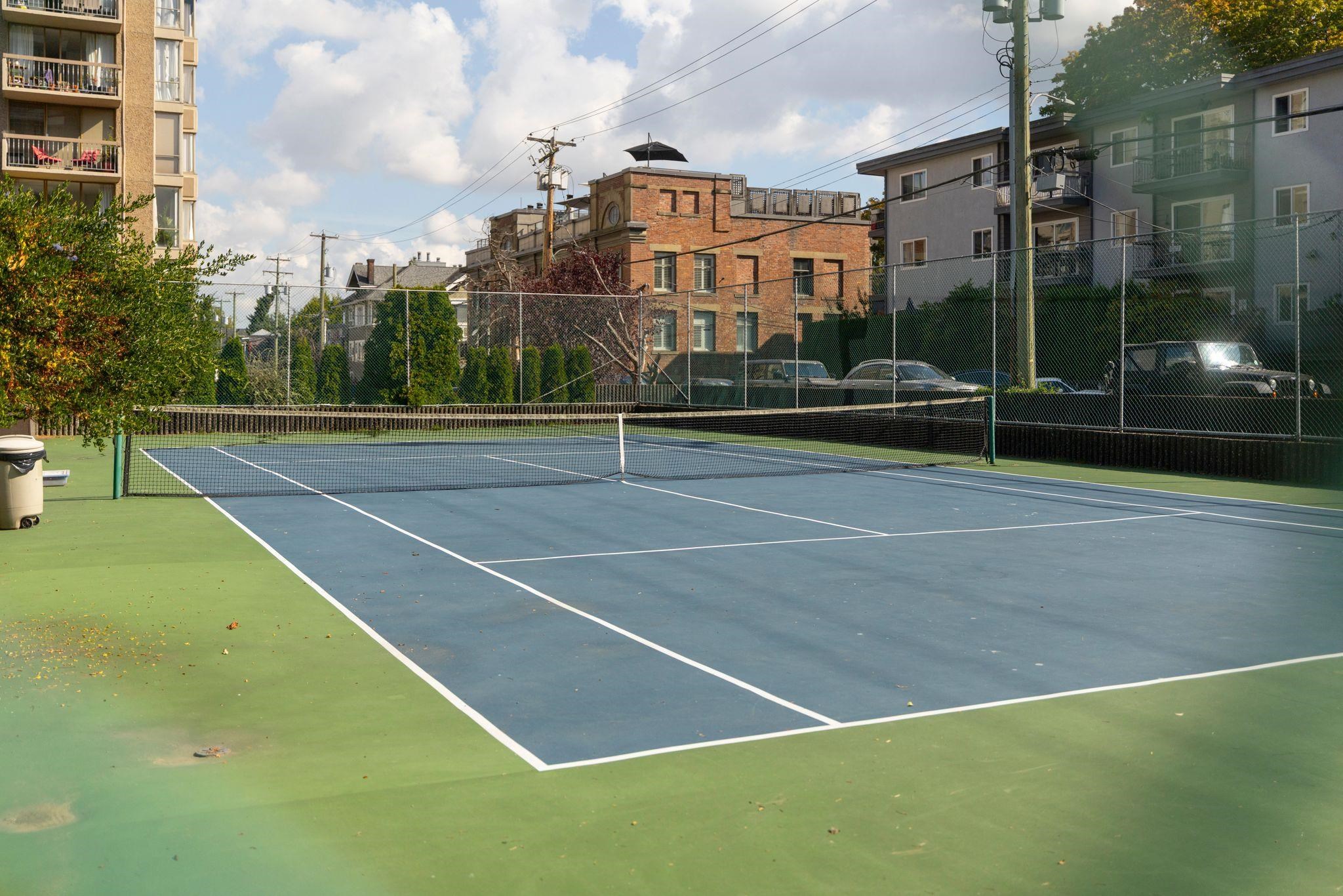 Outdoor tennis court with a blue playing surface, green surround, and chain-link fencing.