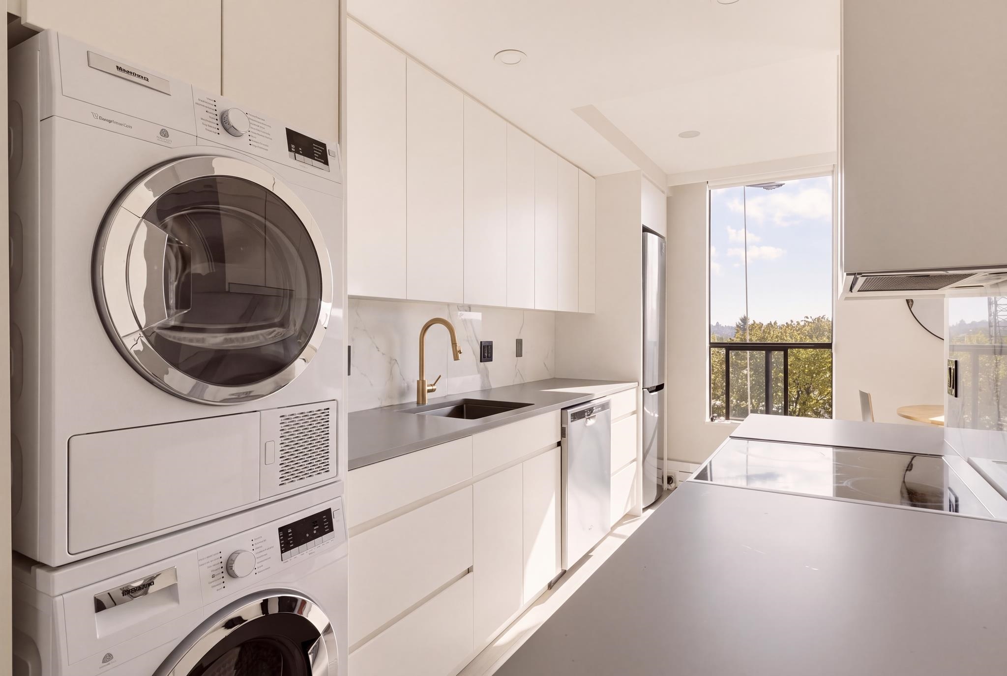 Modern white kitchen with a stacked washer and dryer beside a large window overlooking trees.
