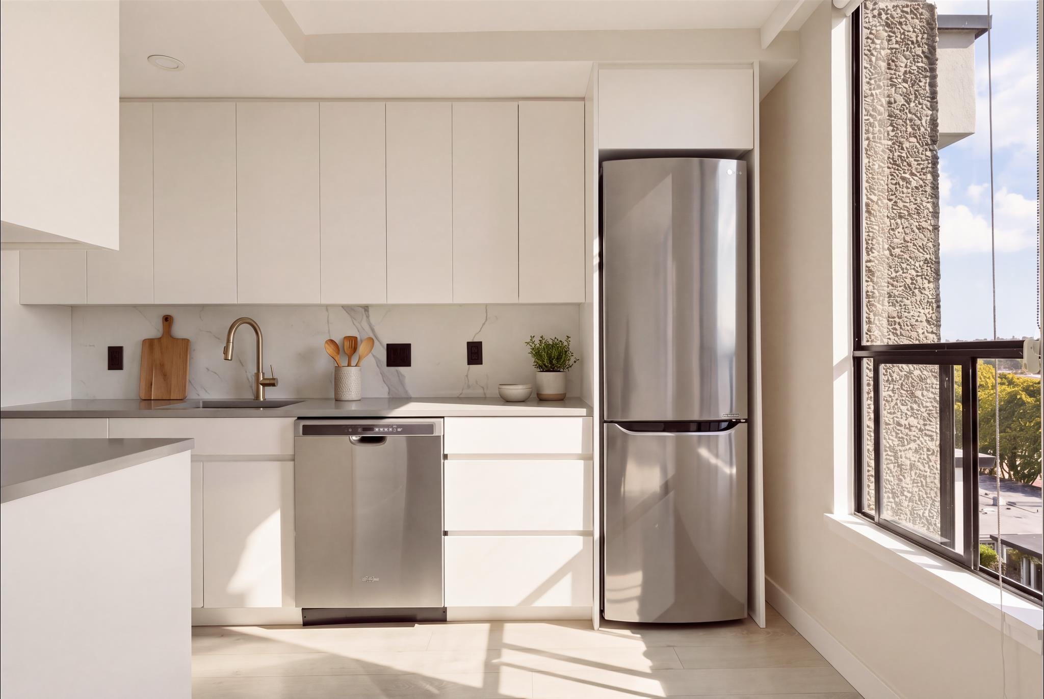 Modern white kitchen with stainless appliances and a large sunlit window beside the refrigerator.